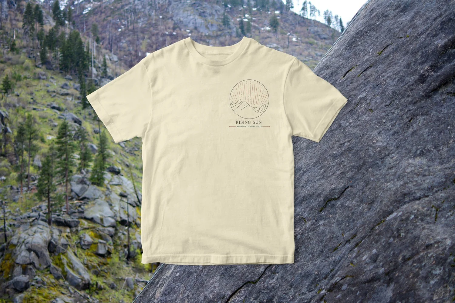 Yellow t-shirt shows circular full color logo on chest. Backgrond: mountainside with pine trees and rocks behind