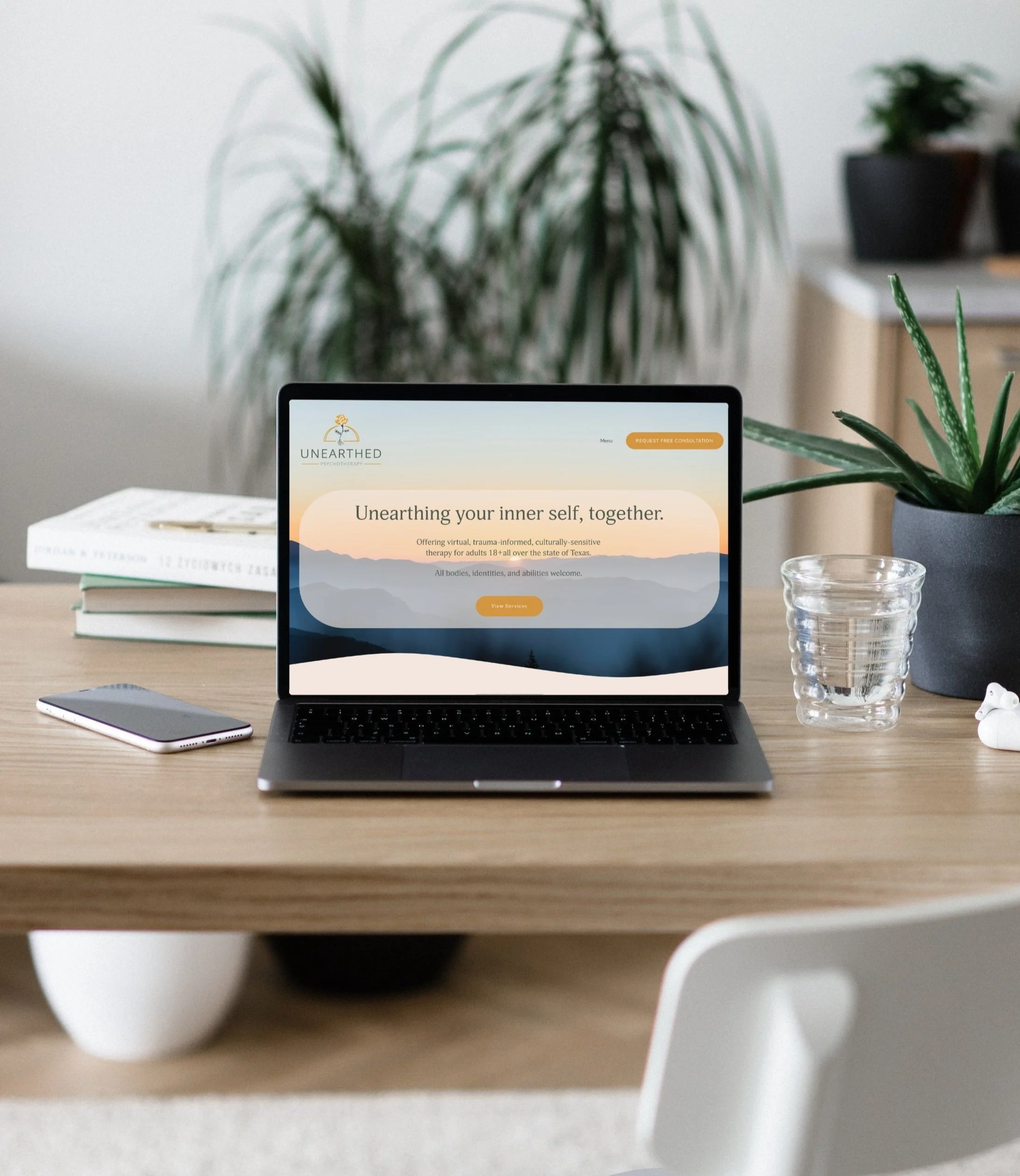 Image Alt Text: Unearthed Psychotherapy's new website is mocked up on a laptop that is placed on a wooden table with a glass of water to the right and a cell phone to the left. A large plant is visible in the background and part of a white chair is v
