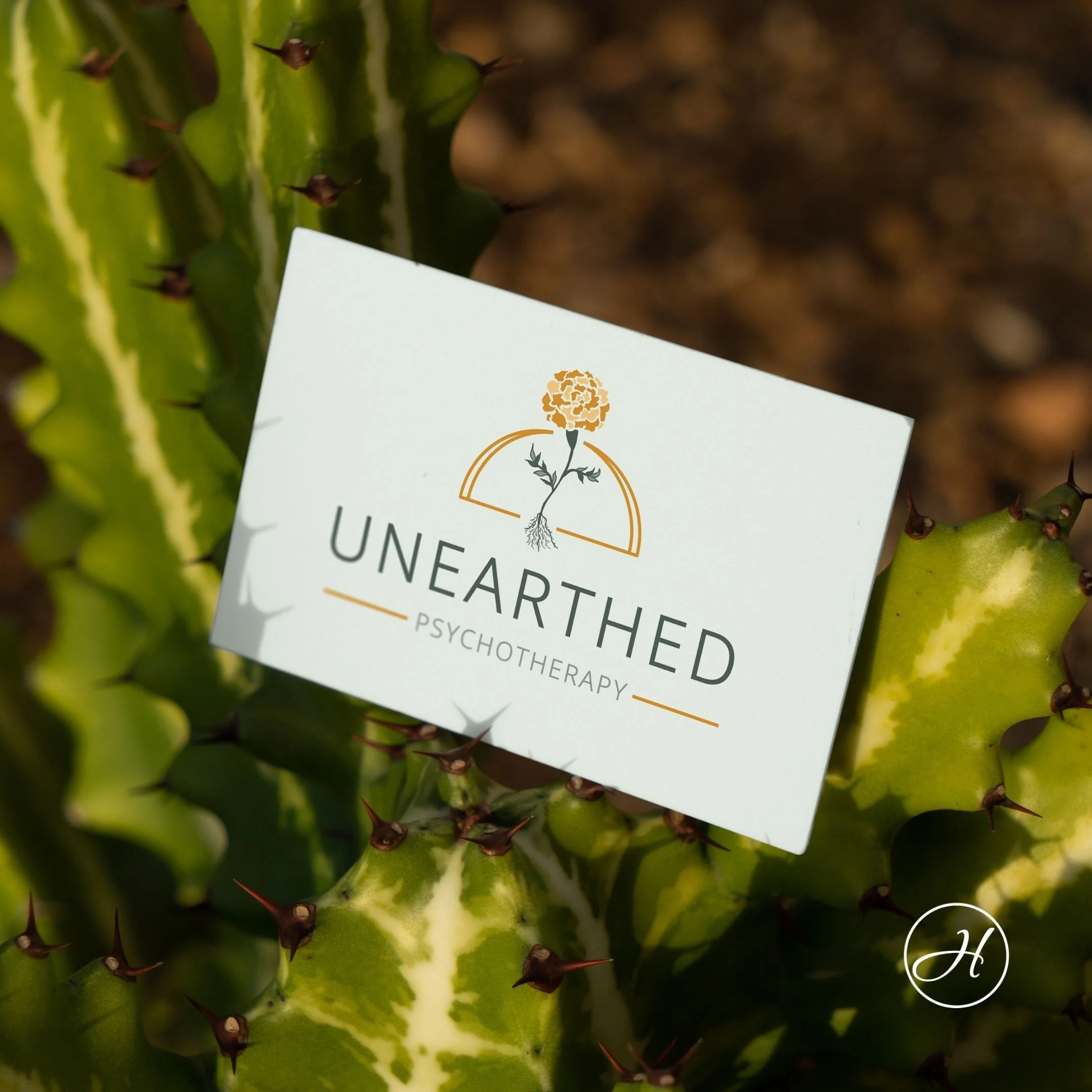 Cactus with business card featuring Unearthed Psychotherapy's logo, an orange marigold with exposed roots in front of an orange semi circle, "Unearthed" underneath, & "Psychotherapy" between orange lines below that.