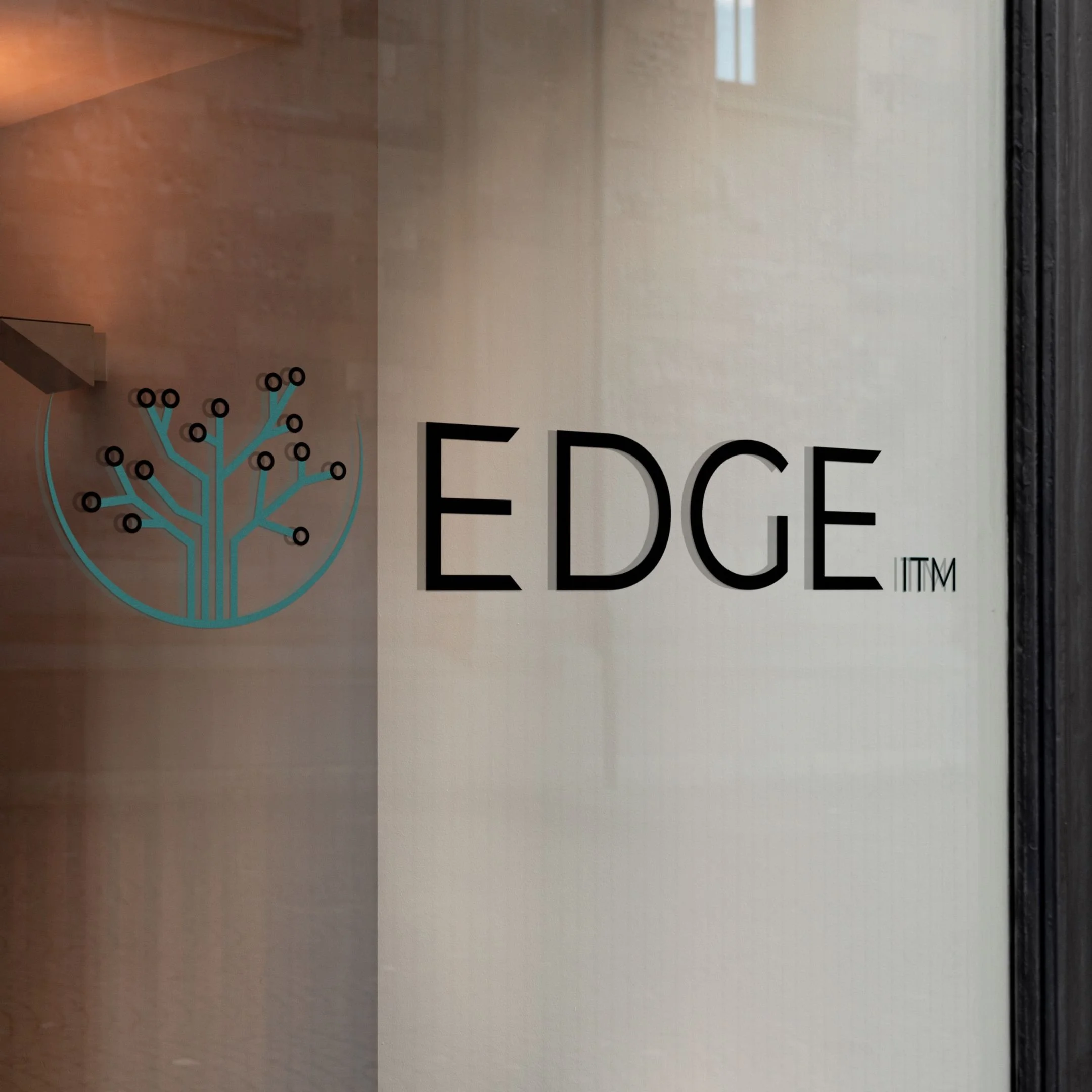 Blue and black circuit-tree design with black "EDGE ITM" to the right, mocked up as window decal, shown at an angle