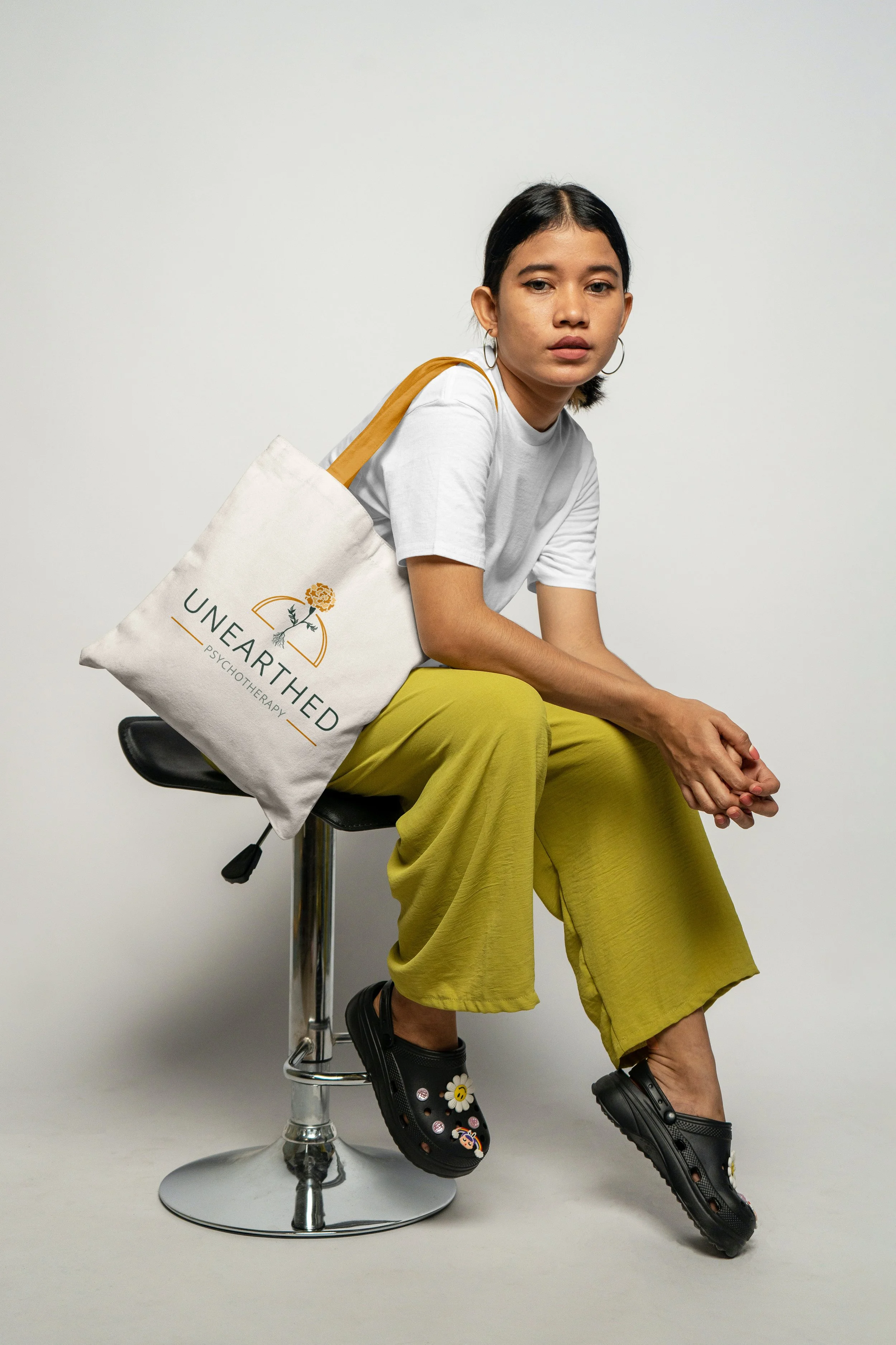 Full color Unearthed Psychotherapy logo is shown on a canvas colored tote bag with yellow/orange straps, on shoulder of seated person with yellow pants and white shirt.
