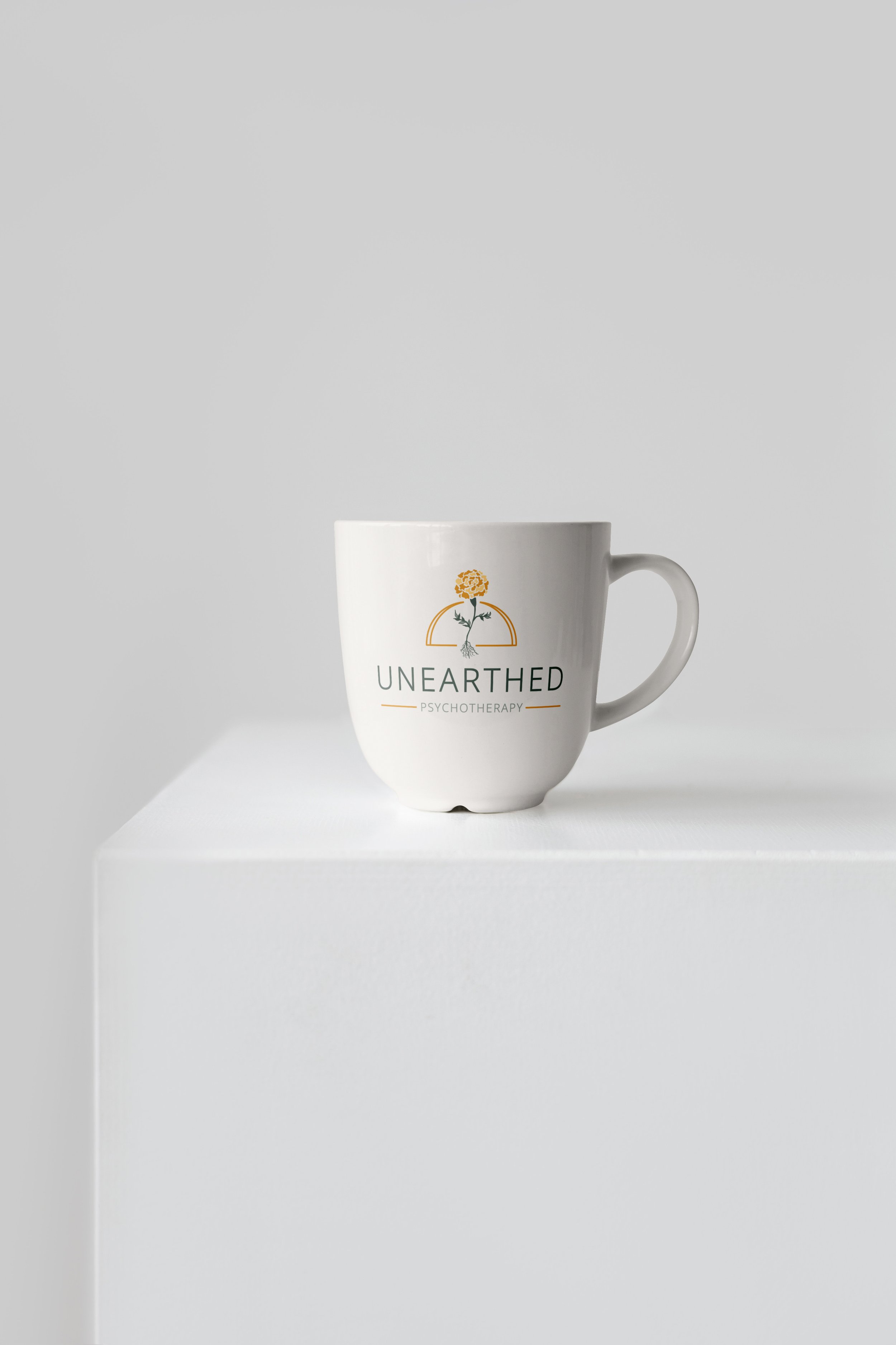The full color Unearthed Psychotherapy logo is mocked up on a white coffee cup, placed on a white display block with a white background