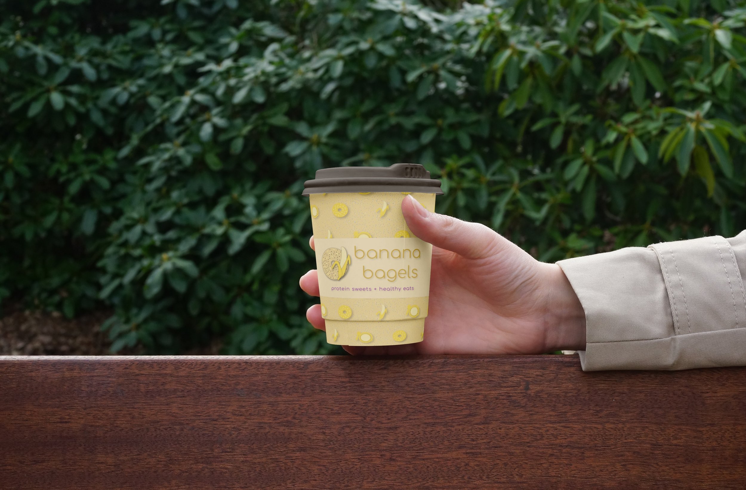 A to-go coffee cup with a brown lid is shown with the previous repeating pattern background and the Banana Bagels logo over top. Held by hand and placed on brown banister with green shrubbery in background.