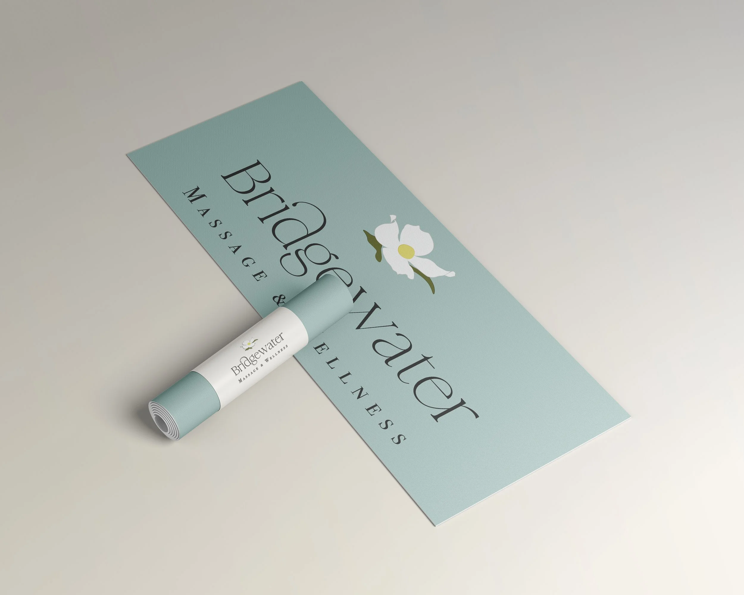 Bridgewater Massage & Wellness' full color logo is shown on light blue background of a rolled out yoga mat. A rolled up yoga mat lies on top with a white label with logo.