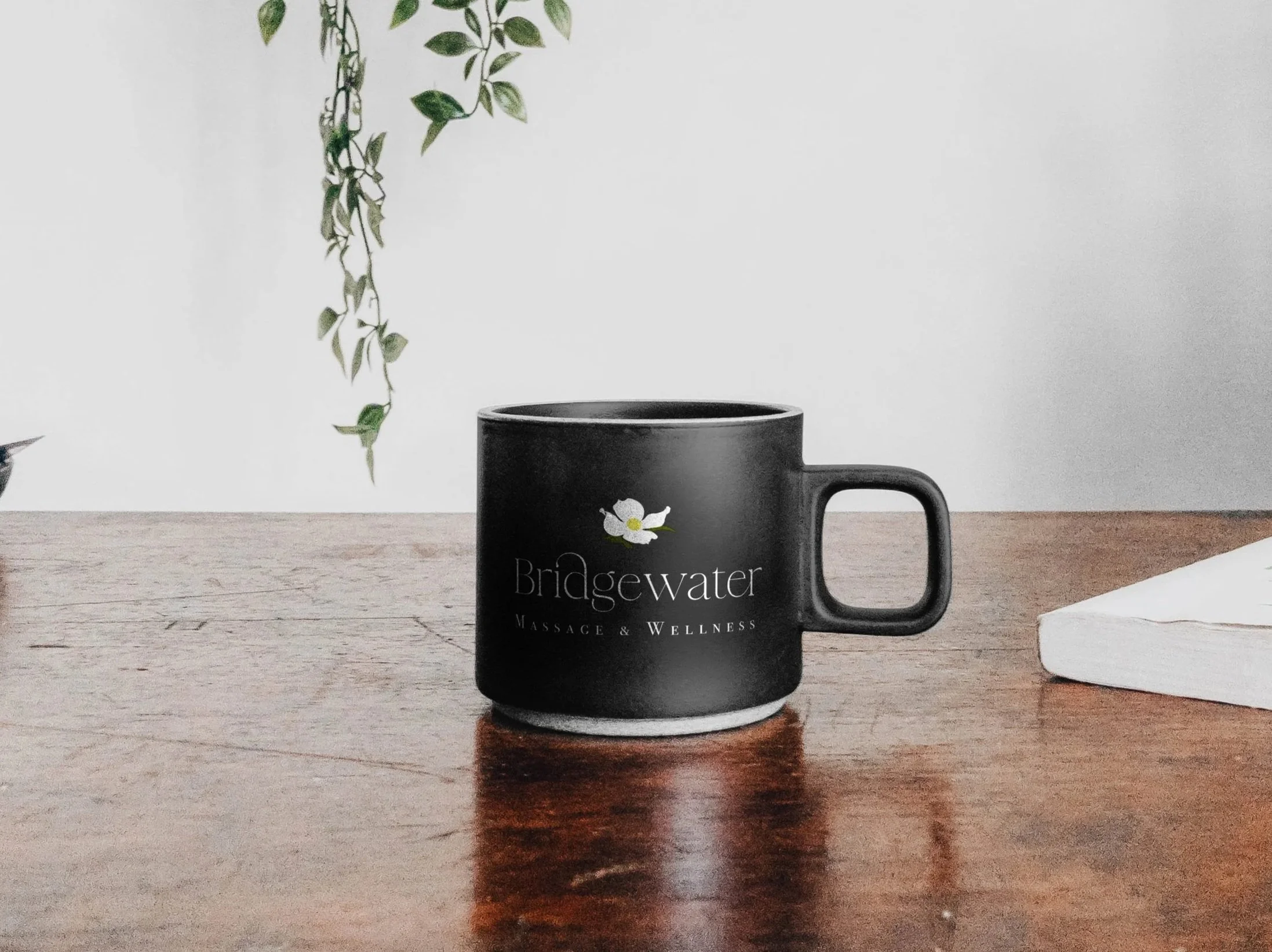 Bridgewater Massage & Wellness' full color logo is shown on a black coffee cup on a wooden table with part of an ivy hanging in background