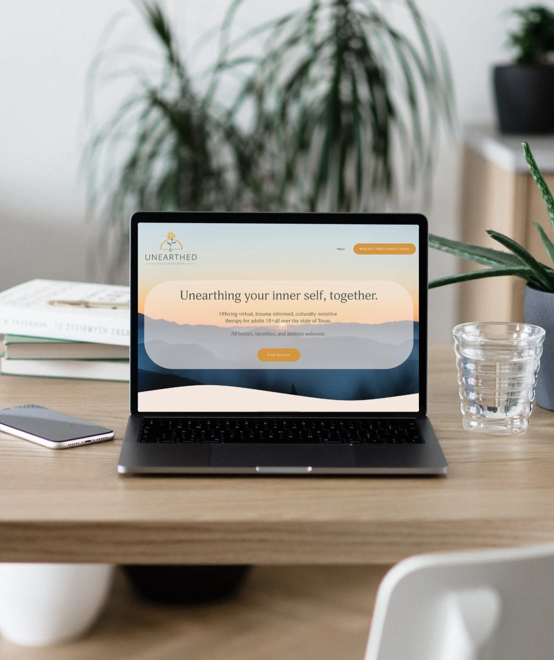 A laptop on a wooden desk displaying a website for psycho therapy called Unearth. The website offers virtual therapy sessions for adults 18+ in Texas. There is a green plant behind the laptop and a glass of water to the right, with a smartphone and a book also on the desk.