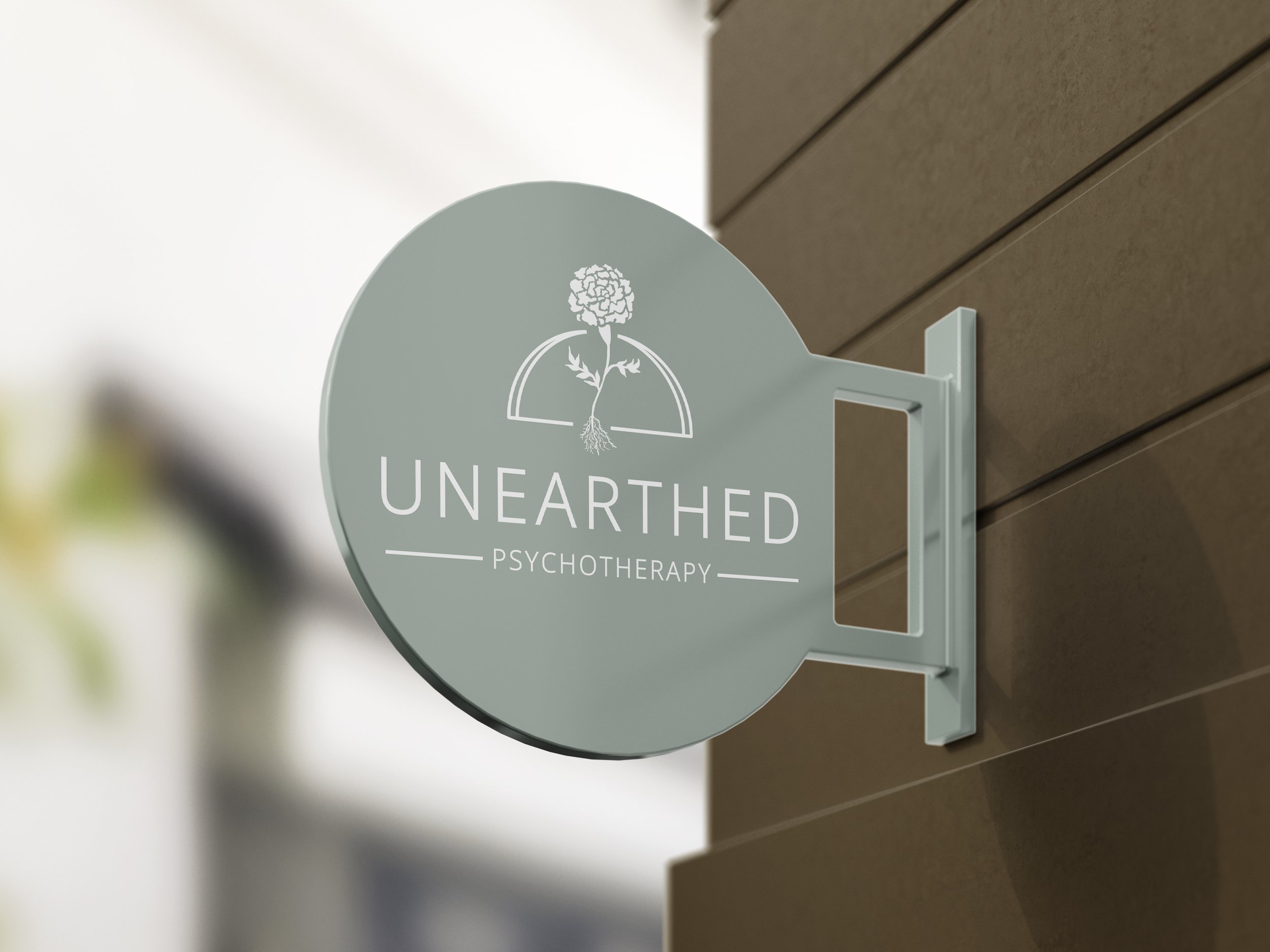 All white Unearthed Psychotherapy logo is mocked up on a light green circular sign, placed on the side of a building with brown paneling