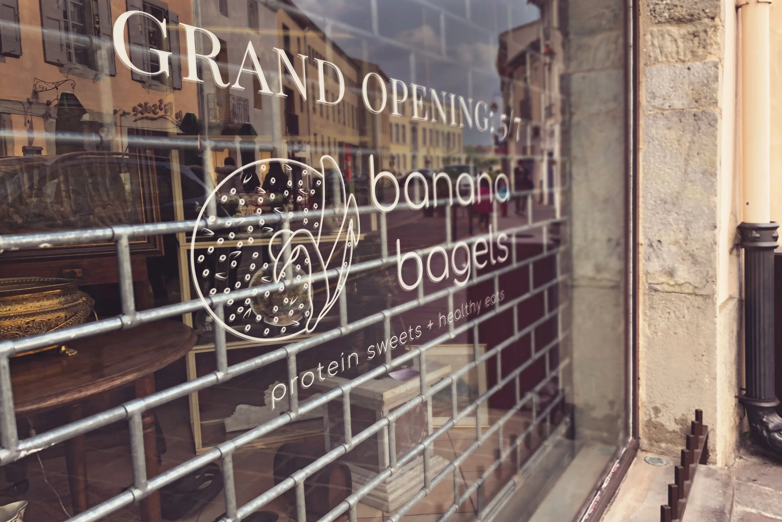 All white logo is mocked up on window, shown at an angle, with "GRAND OPENING 5/7" above.