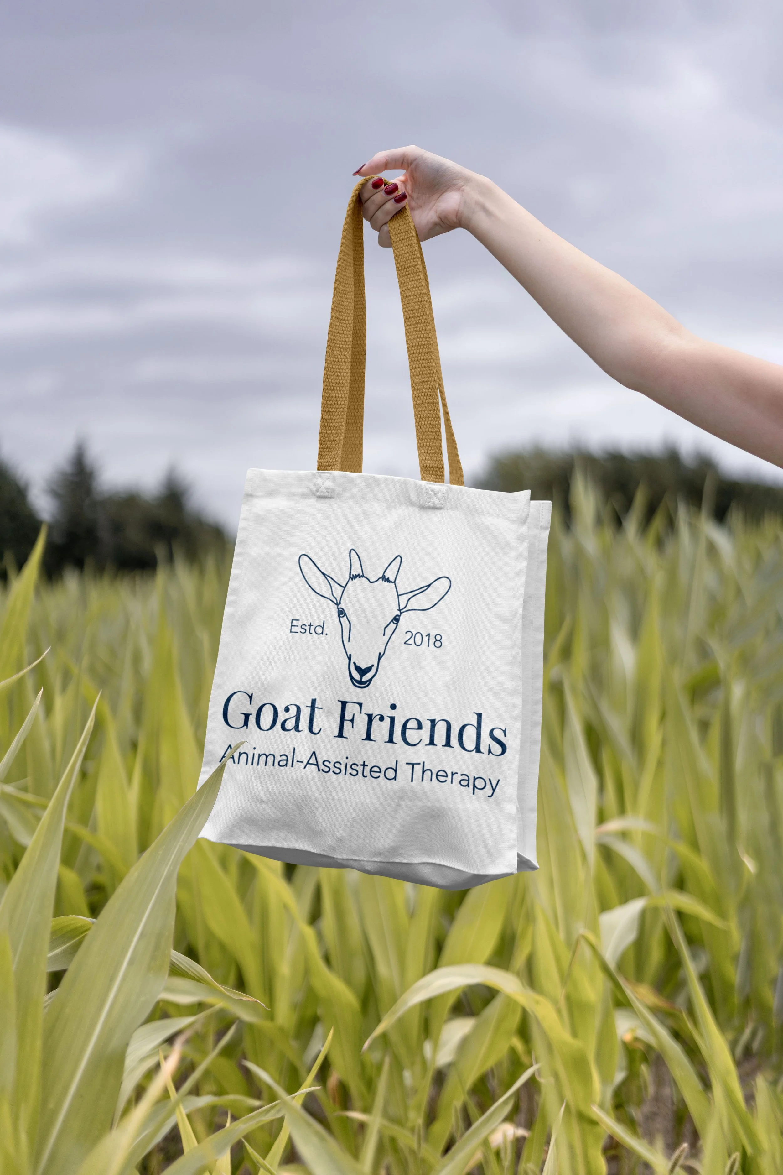 All blue Goat Friends logo is mocked up on a canvas bag with yellow straps, shown in a field of green corn leaves with a blue/gray sky and dark green trees on the horizon.