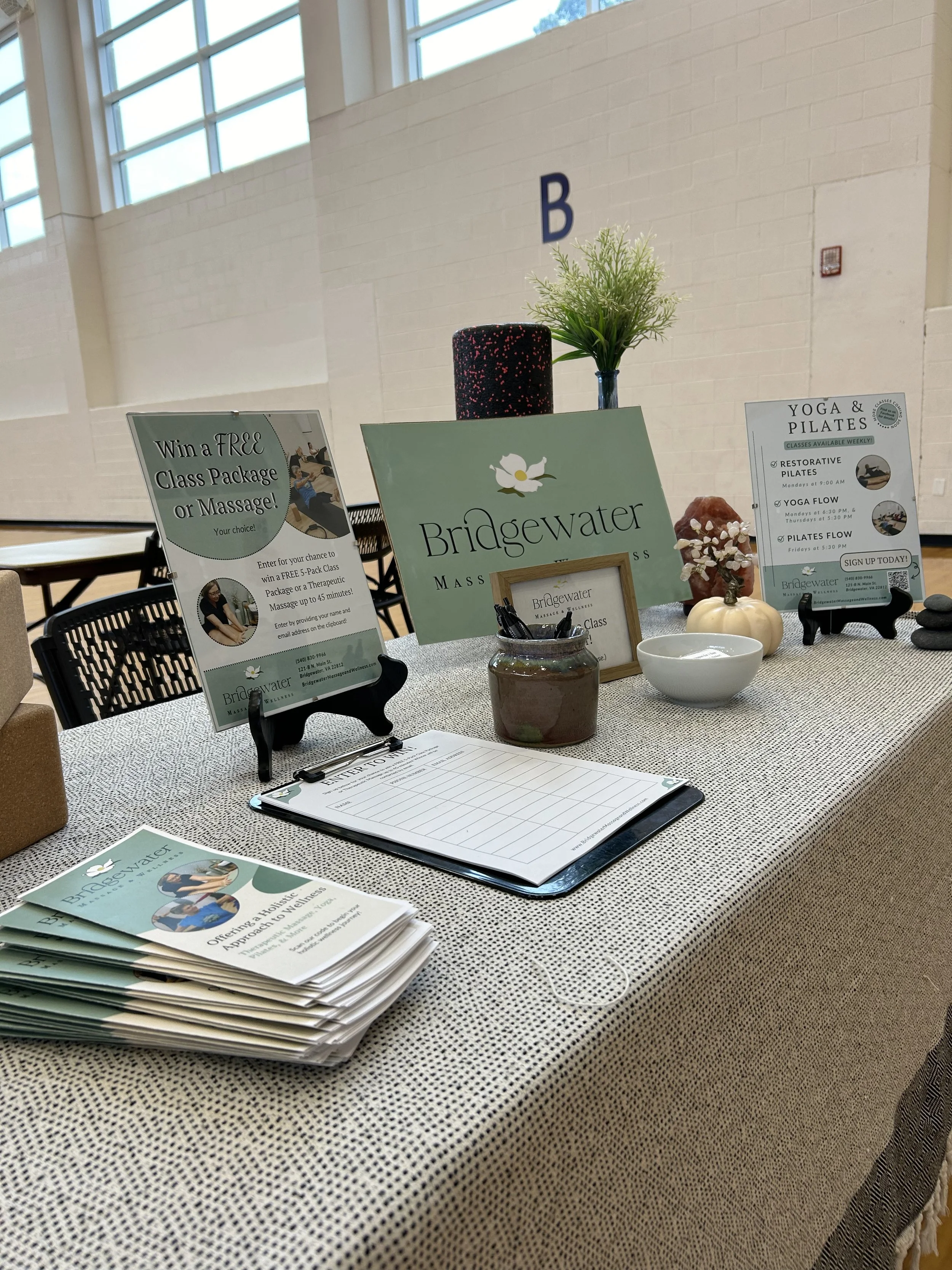 A gray clothed table features marketing materials designed by Heartleaf, including a giveaway sign, brochures, signup sheet, and class schedule. Also shows various table decor.