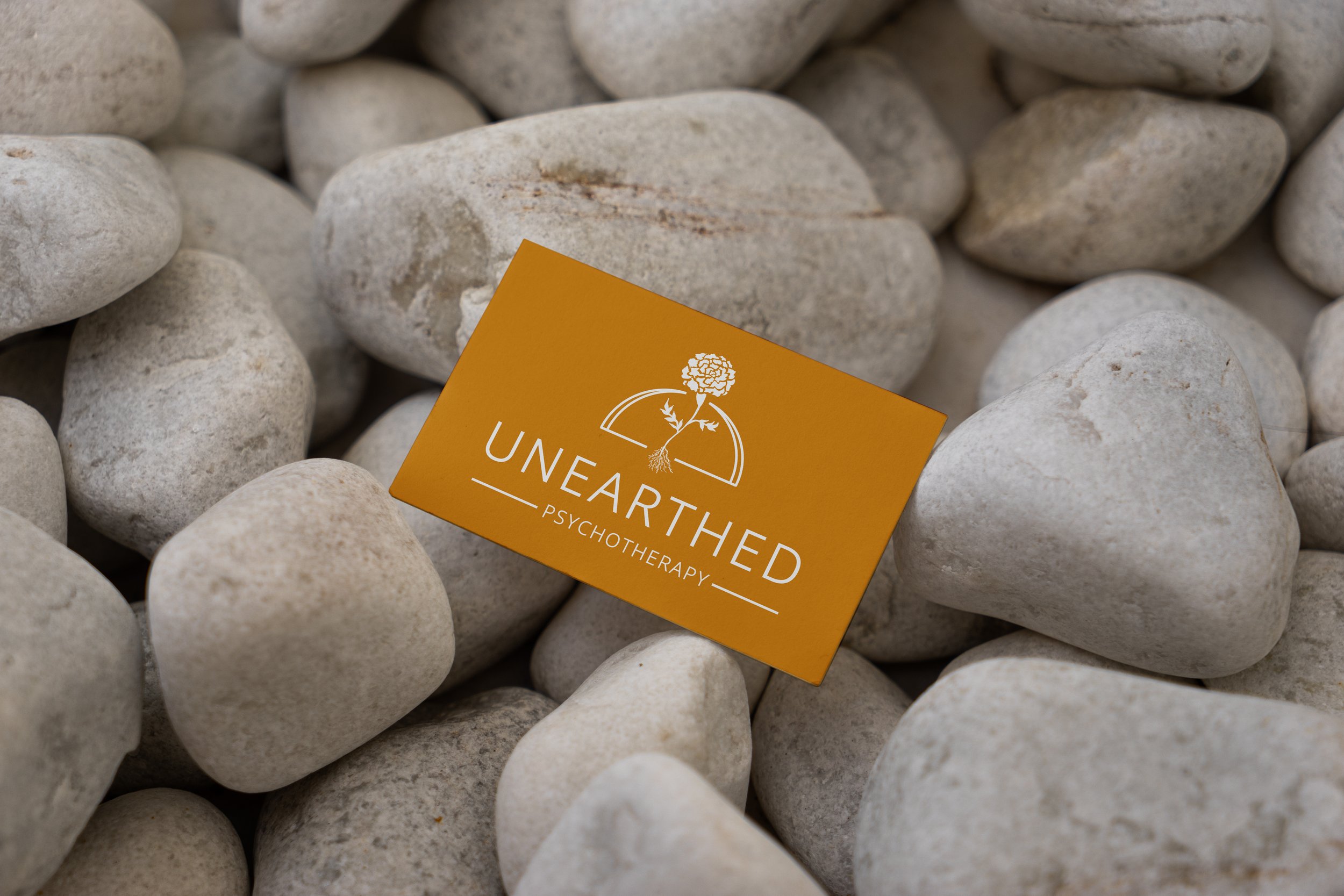 White Unearthed Psychotherapy logo on a dark yellow/orange business card, placed on a background of white river rocks