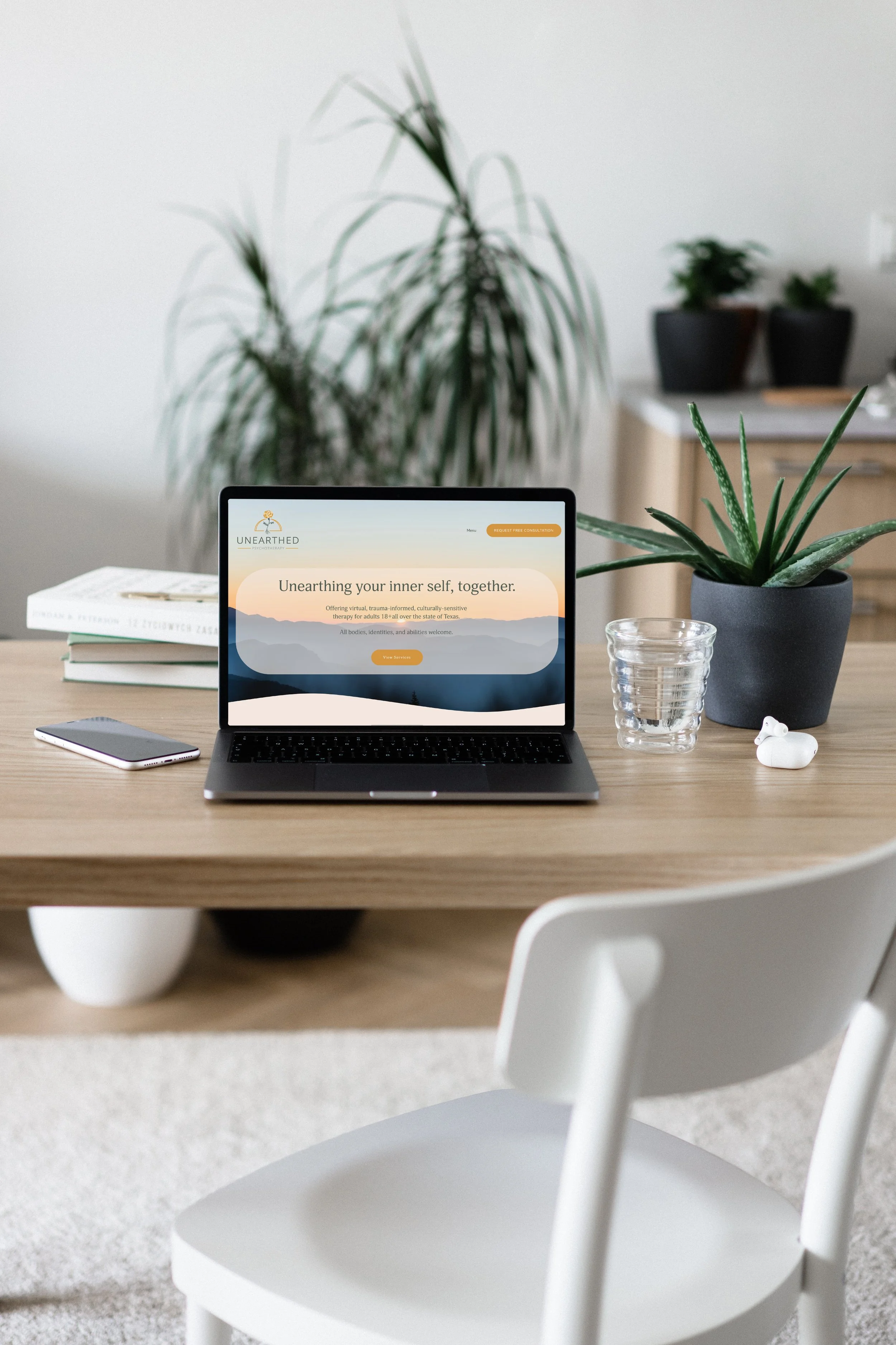 Unearthed Psychotherapy's new home is mocked on a laptop, placed on a wooden table with plants behind, a water glass to the right, & a cell phone to the right.