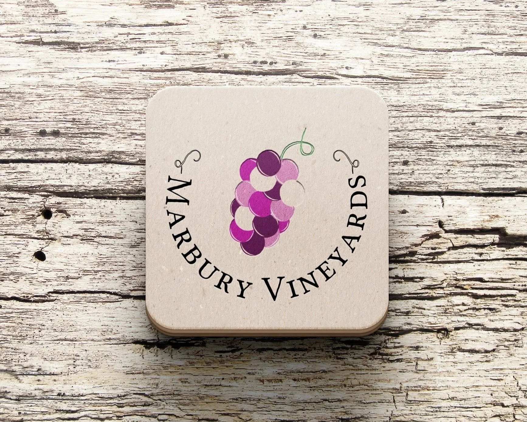 A coaster with Marbury Vineyards' logo and grapes with purple shades in the center placed on a gray wooden table
