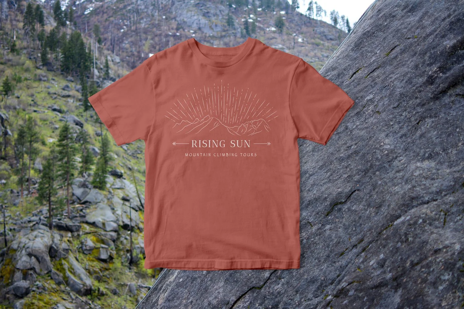 Red t-shirt shows white logo centered on shirt. Backgrond: mountainside with pine trees and rocks behind