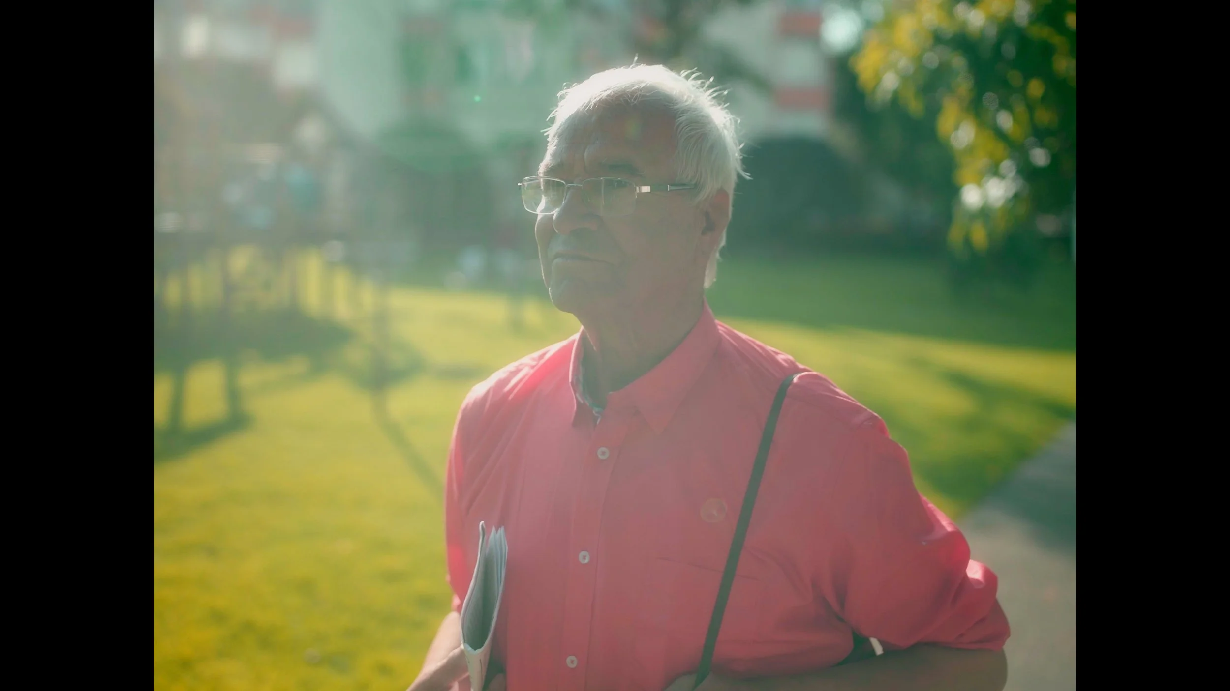 Cinematography, dirección de fotografía, DoP, cortometraje. Anciano con gafas y camisa rosa, sosteniendo algunos papeles, en un parque con árboles y sol en el fondo.