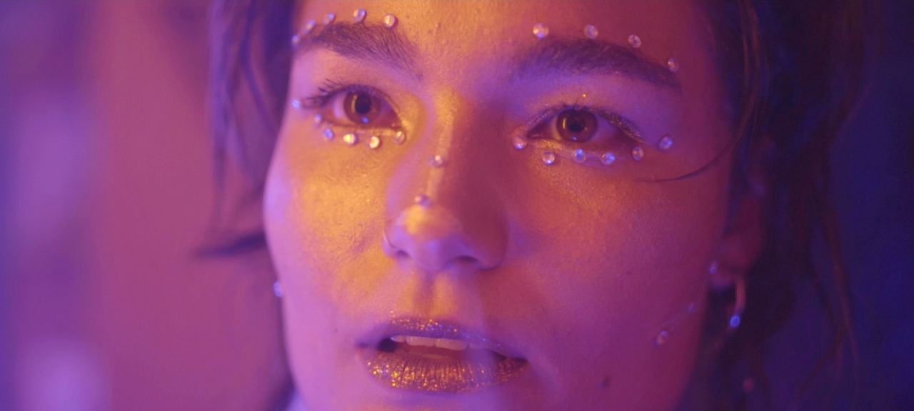 Retrato de una persona con maquillaje brillante y decoraciones de perlas en la cara, con iluminación de colores vibrantes.