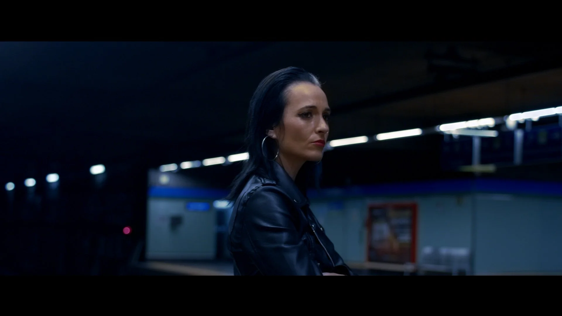 Mujer de cabello negro y maquillaje oscuro, vestida con chaqueta de cuero, en una estación de metro con fondo oscuro y luces brillantes.