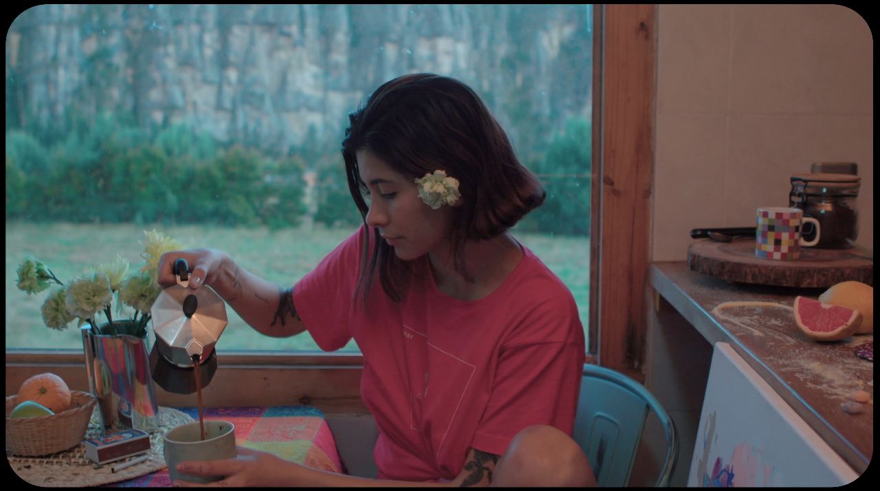 Mujer con flores en el cabello sirviendo café en una taza dentro de una cocina junto a un florero con flores, con vista a un campo y montañas a través de la ventana.