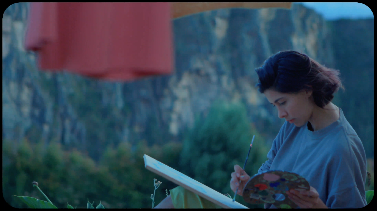 Una mujer joven pintando en un cuaderno al aire libre, con un fondo de montaña y vegetación.