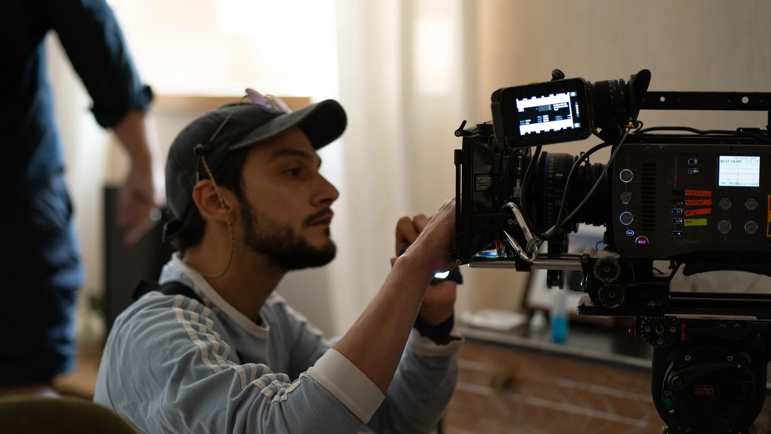 Hombre ajustando una cámara de filmación en un set de grabación, con fondo difuso y otra persona parcialmente visible.