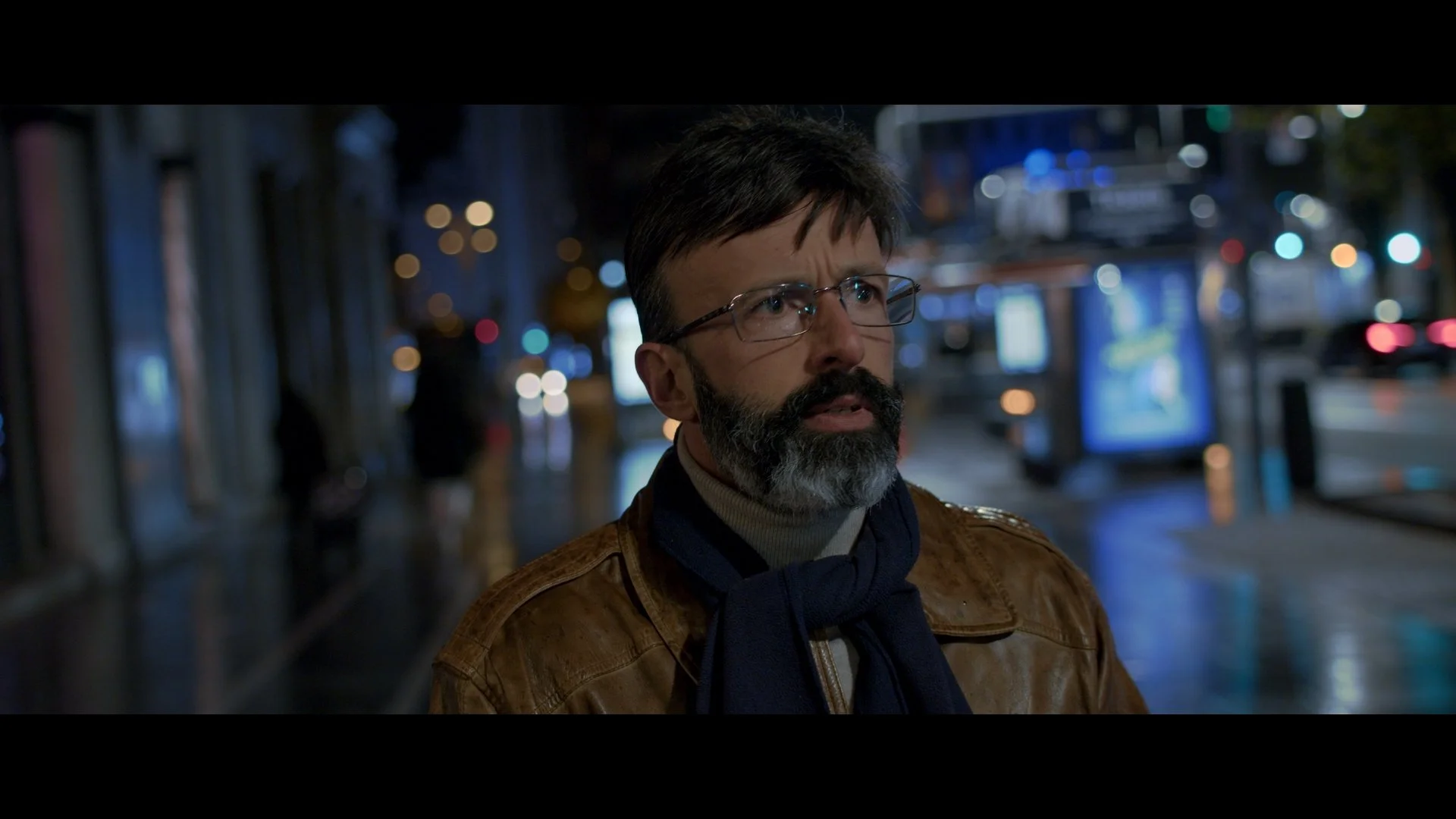 Hombre con barba y gafas de noche en la calle, con luces de neón y anuncios de teléfono en el fondo.