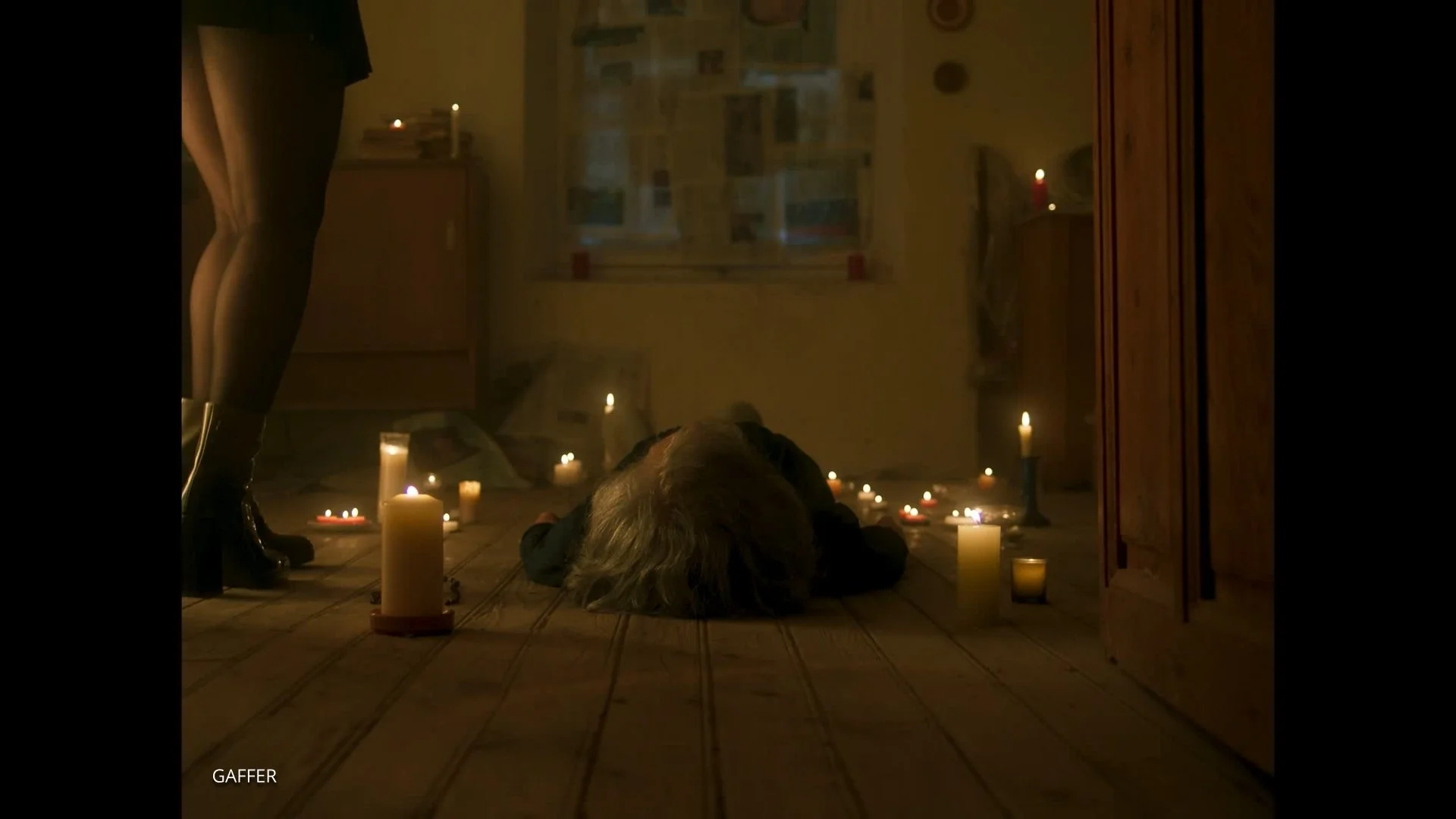 Una persona acostada en el suelo de una habitación oscura, rodeada de velas encendidas en diferentes alturas y tamaños, con una ventana al fondo y un armario de madera a la derecha, en un ambiente tenue y misterioso.