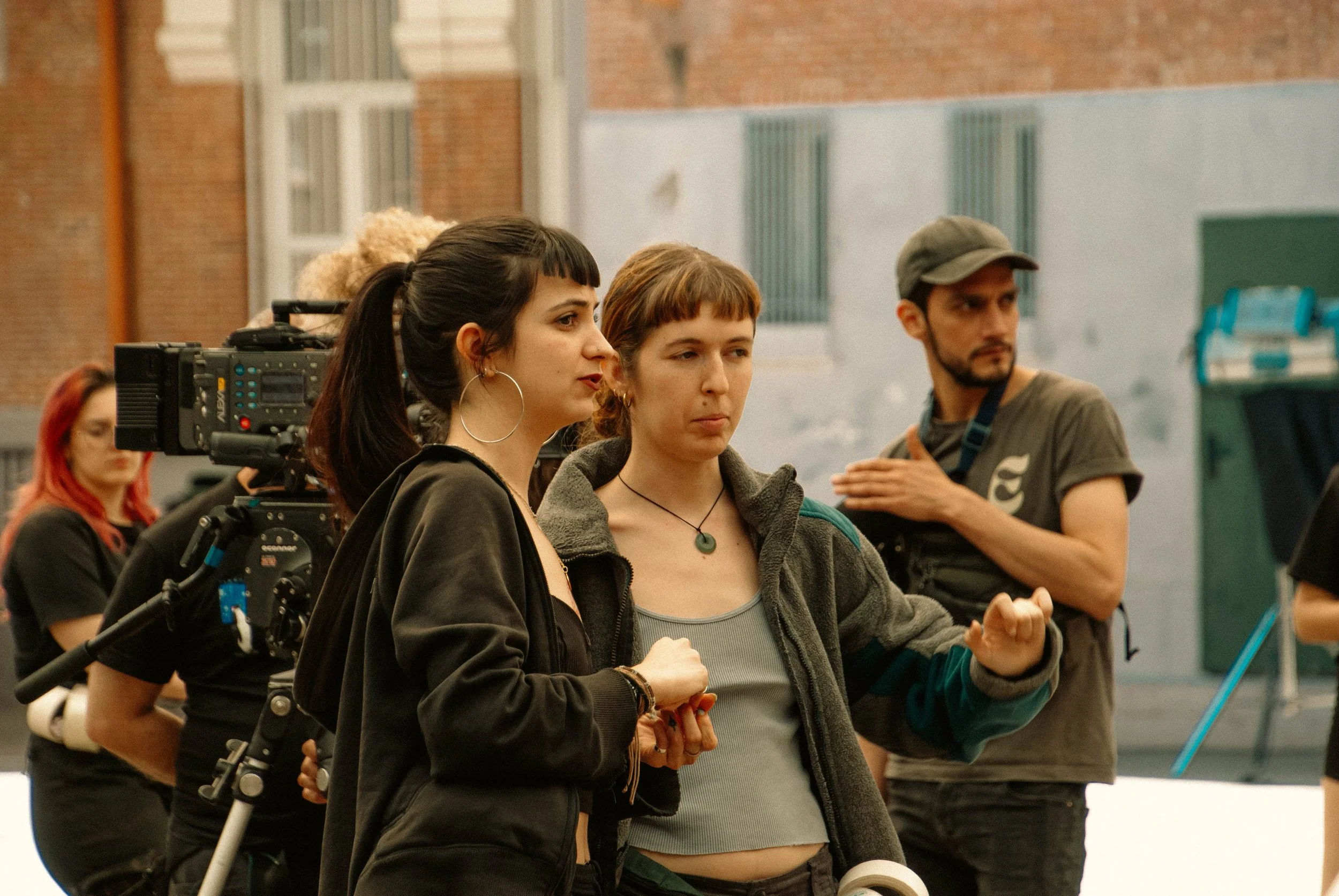 Grupo de personas en un set de filmación, con cámara y equipo de grabación, en una calle con paredes de ladrillo y edificios.