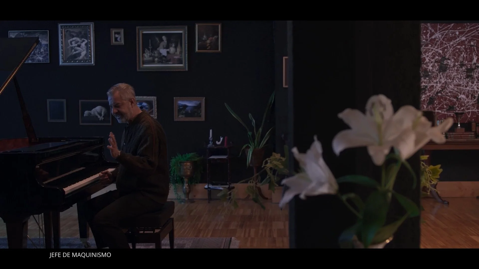 Hombre tocando piano en una habitación con paredes negras y cuadros decorativos, flores blancas en primer plano a la derecha, y espacio elegante y artístico.