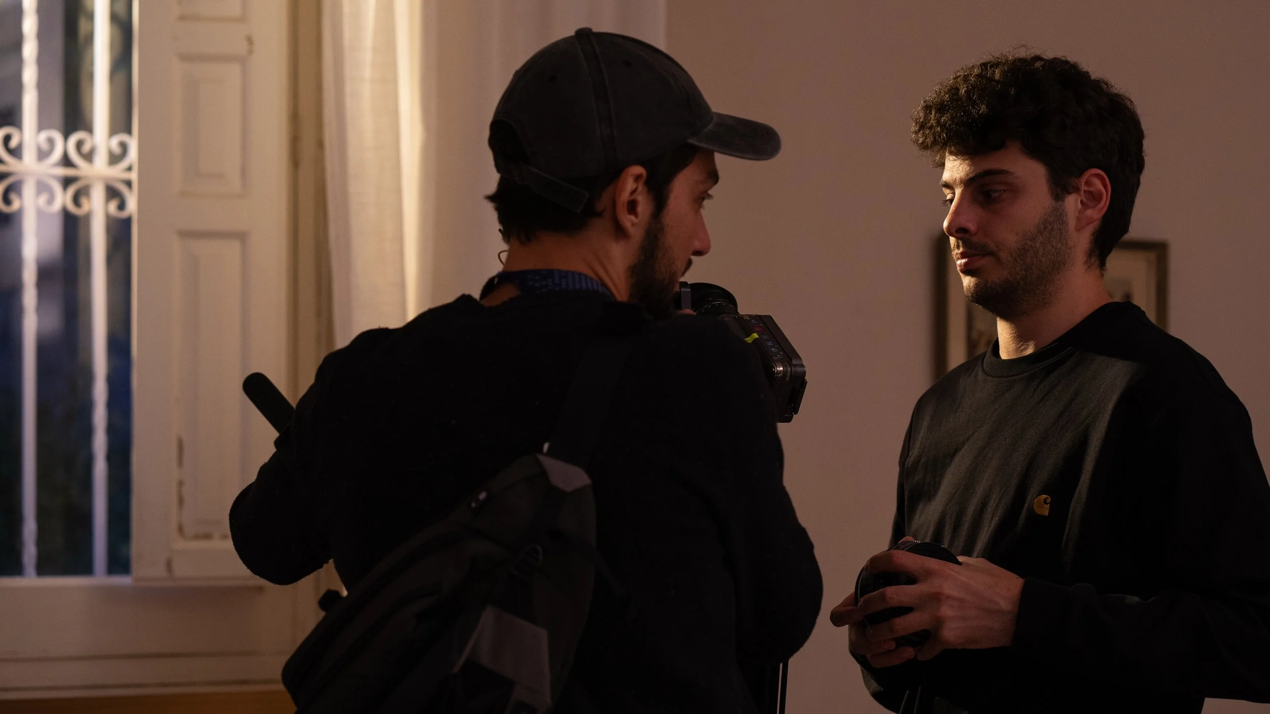 Dos jóvenes están en una conversación en un interior, uno de ellos lleva gorra y mochila, el otro sostiene un objeto en las manos.