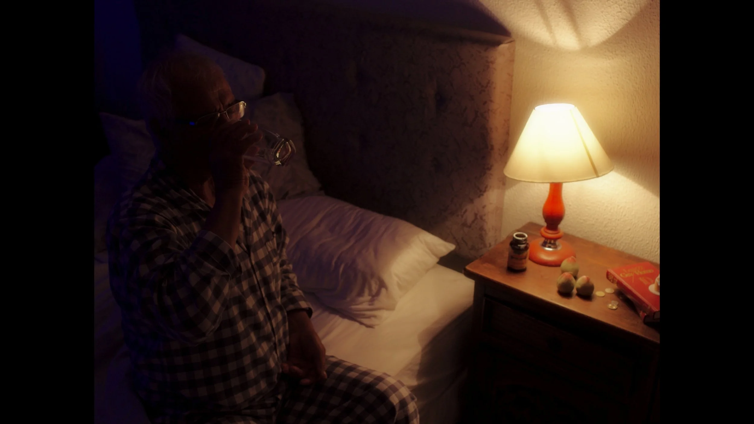 Cinematography, dirección de fotografía, DoP, cortometraje. Anciano tomando agua en la cama, en una habitación con una lámpara encendida sobre una mesita con medicamentos, monedas, un libro y unas piedras decorativas.