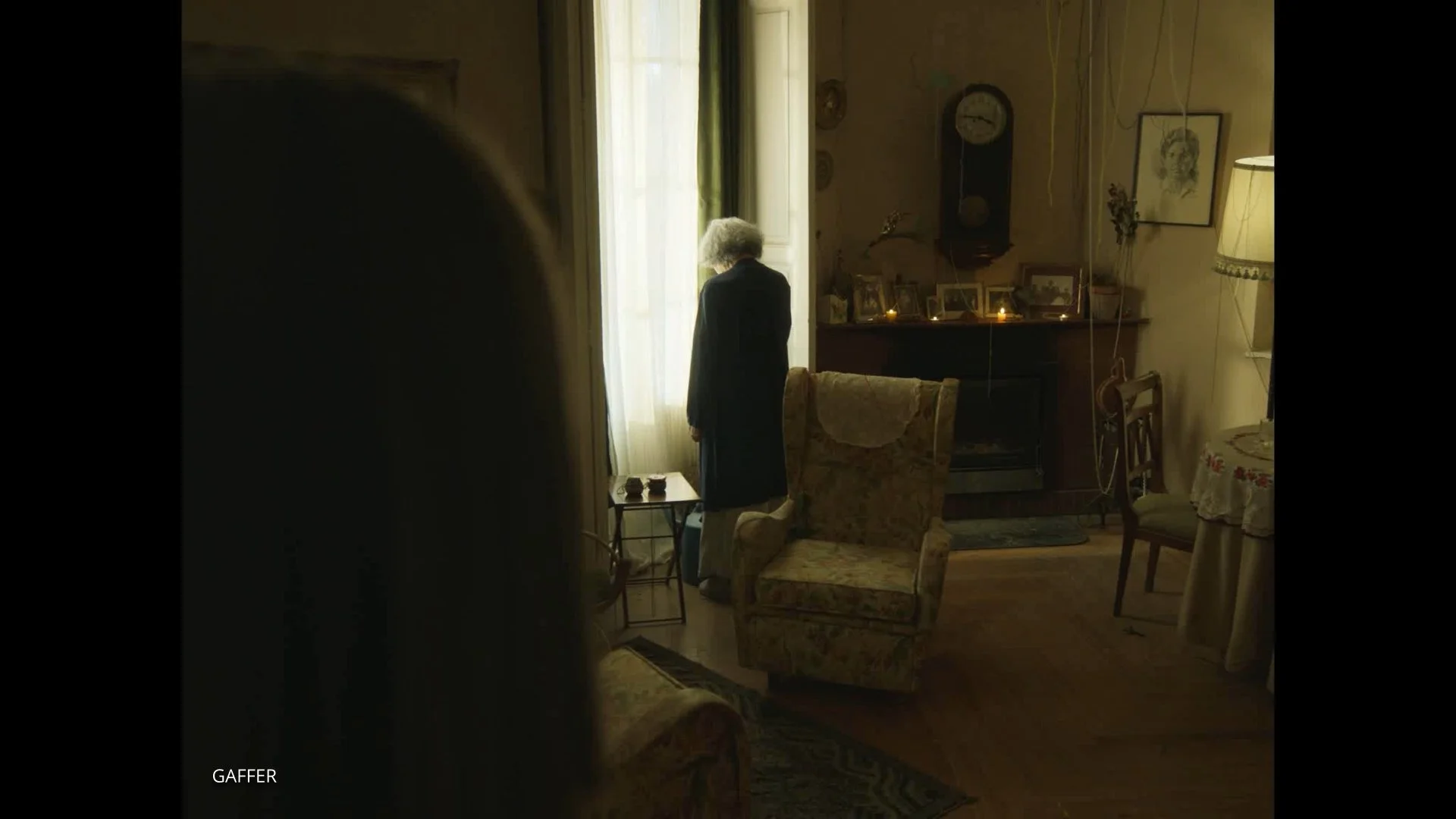 Una mujer mayor de espaldas, de cabello gris, usando un abrigo oscuro, mira por la ventana en una sala de estar acogedora con decoración vintage y una chimenea con fotografías y velas encendidas.