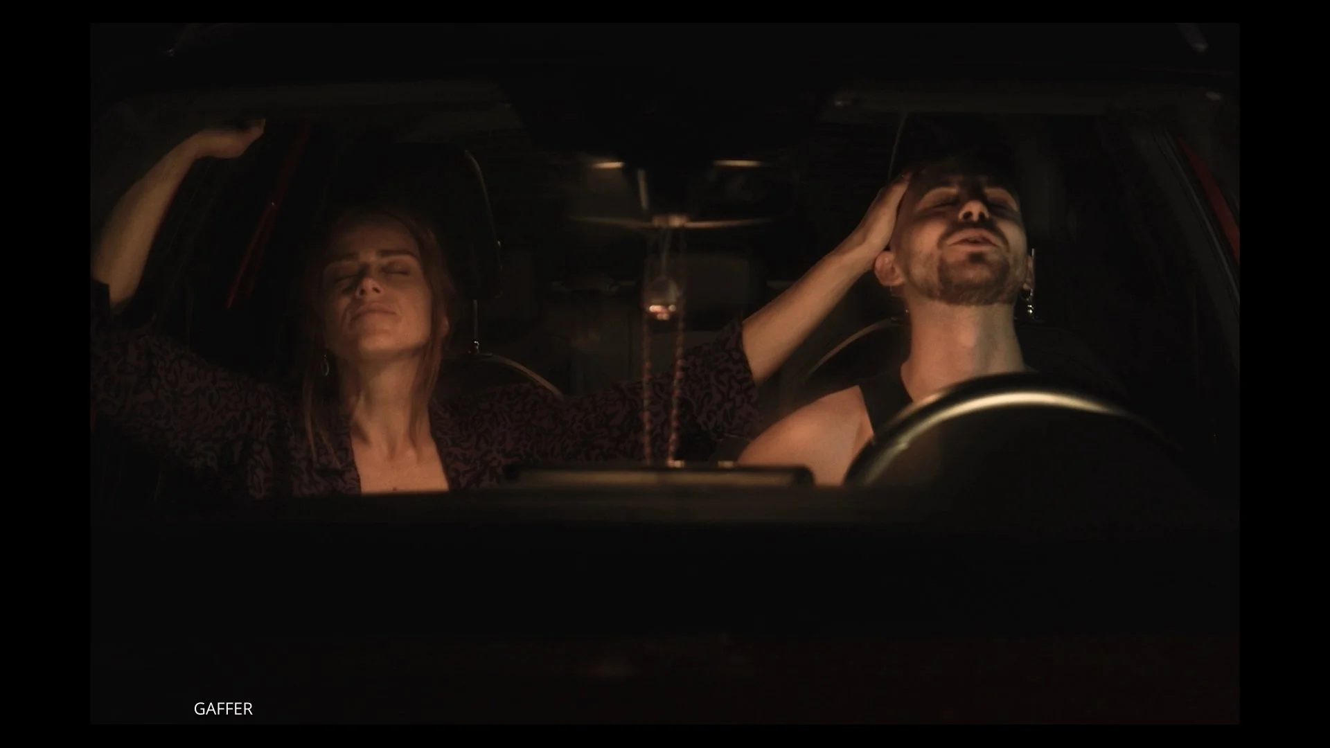 Una mujer y un hombre en el interior de un coche en la oscuridad, ambos con los ojos cerrados y tocándose la cabeza, parece que están en un momento de introspección o meditación.
