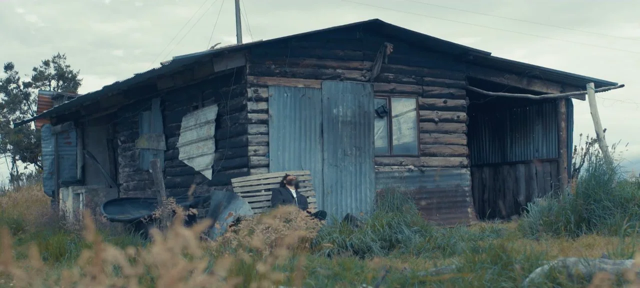 Una casa vieja y deteriorada construida con madera y latas, en un campo con pasto alto y una persona sentada en un banco con árboles en el fondo.
