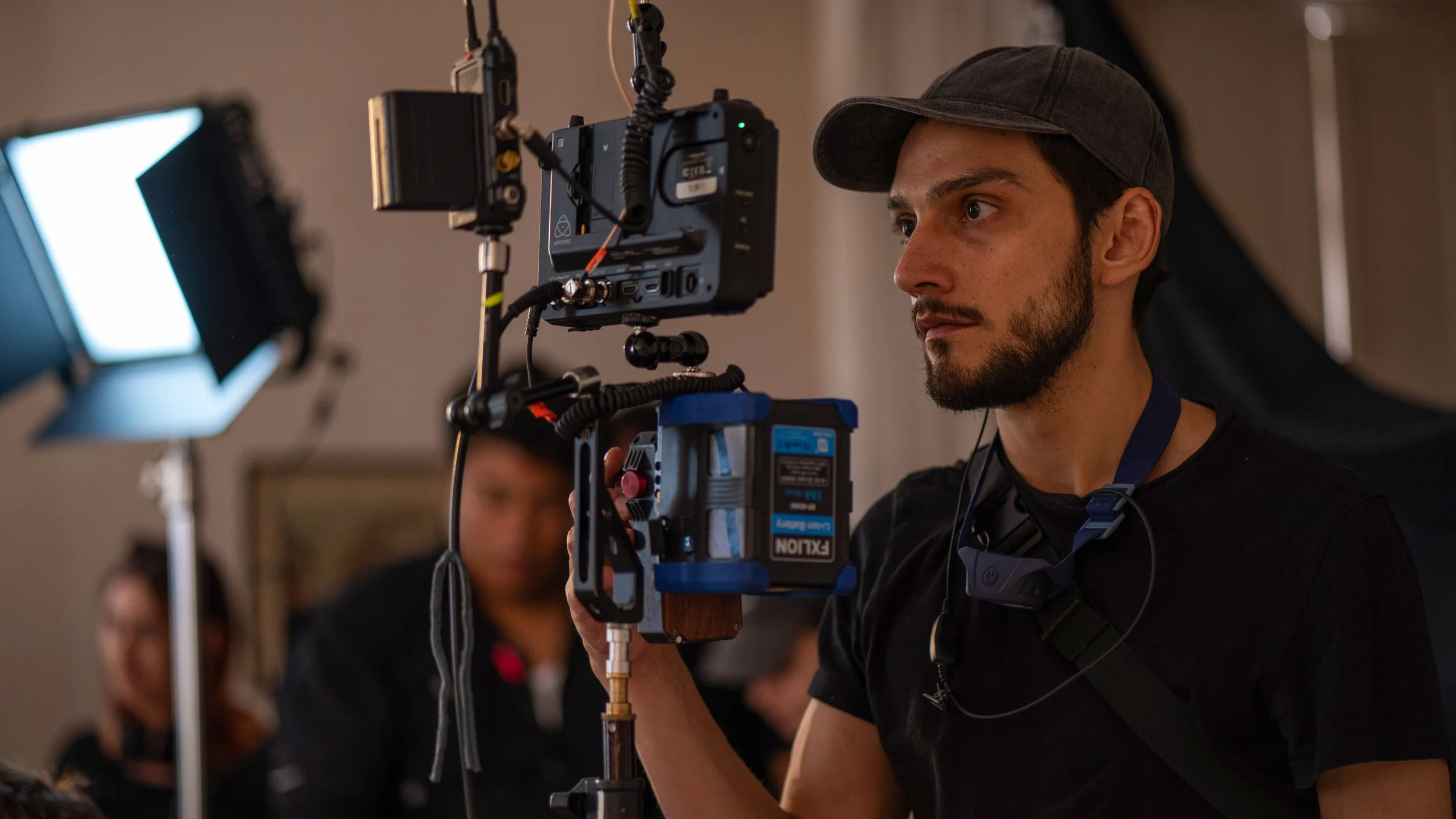 Hombre en un set de filmación ajustando una cámara profesional, con luces y personas trabajando en el fondo.