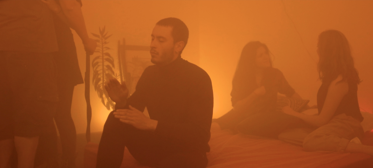 Cuatro personas en una habitación con iluminación anaranjada; un hombre parece estar orando o meditando, y dos mujeres están sentadas en la cama hablando, mientras otra persona en el fondo está de pie.