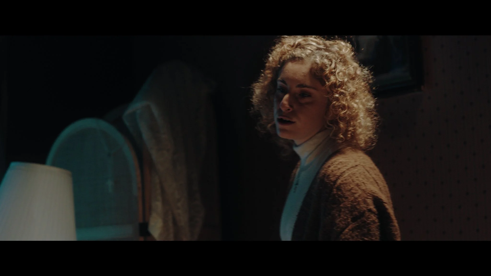 Mujer con cabello rizado y expresión de sorpresa en una habitación oscura con lámpara y espejo vintage.