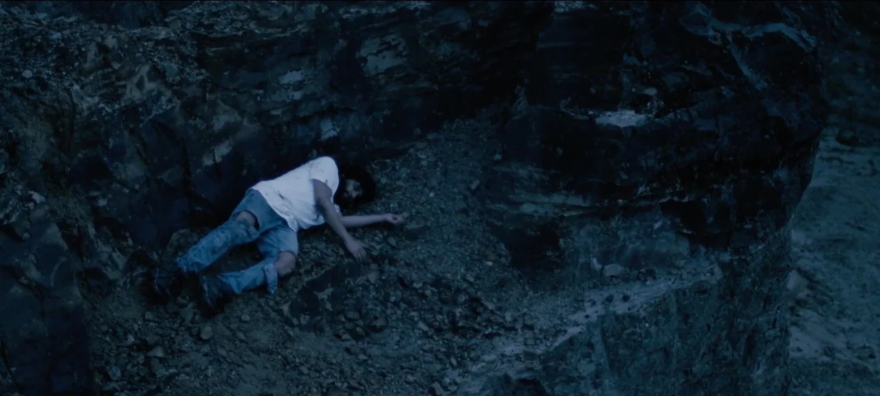 Joven mujer dormida en una superficie rocosa, parcialmente acostada de lado, en un entorno natural con rocas y tierra.