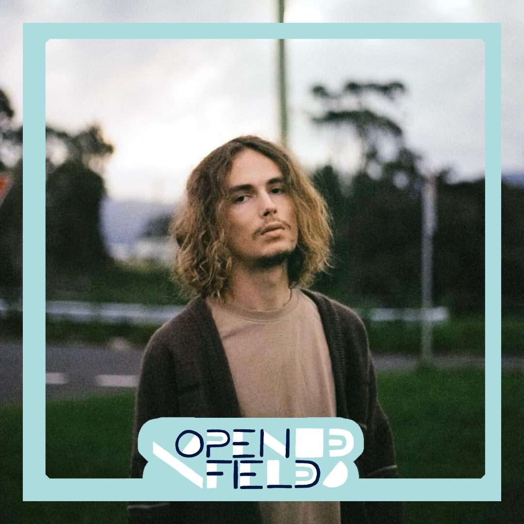 Person with long curly hair wearing a brown jacket standing outdoors with blurred greenery in the background, surrounded by a graphic border with the text 'OPEN FIELD.'