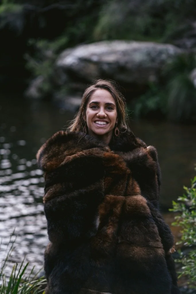 Woman wrapped in a brown fur shawl smiling by a riverside with trees and rocks in the background.