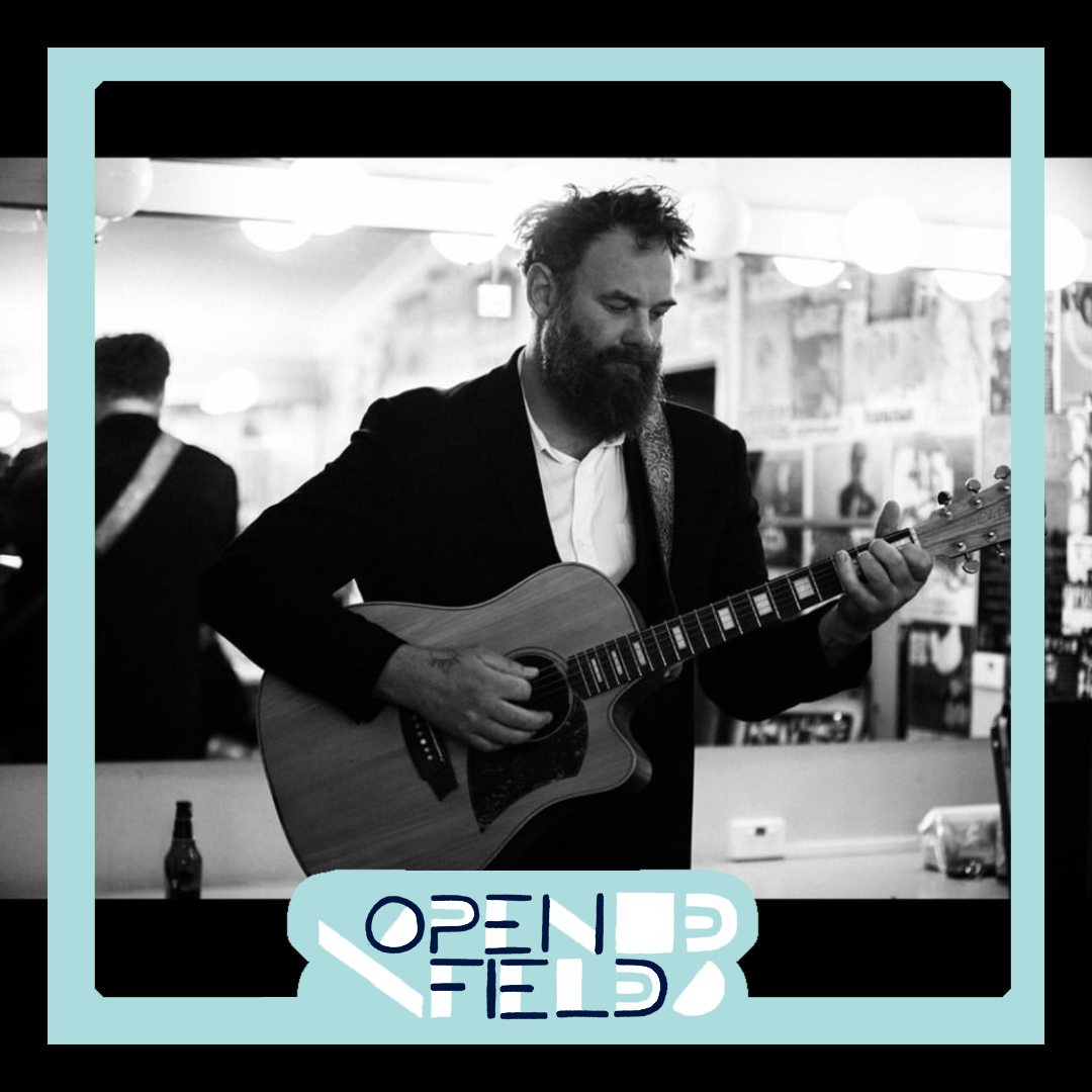 Man playing acoustic guitar in dressing room, surrounded by music posters, with "Open Field" text overlay