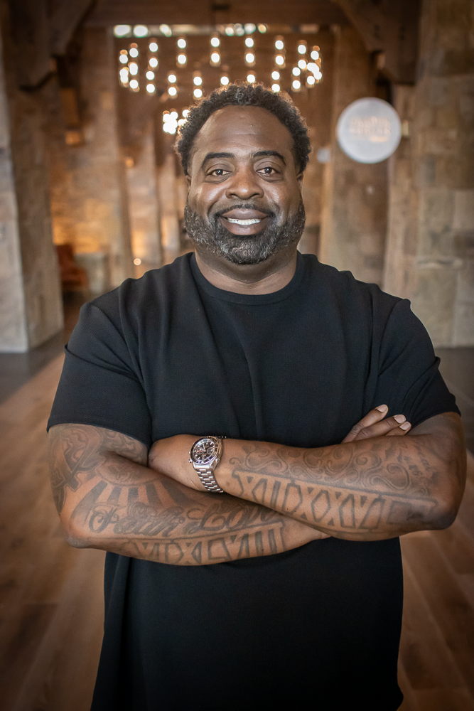 A picture of Damion Wright smiling with facial hair and tattoos on his arms, standing with arms crossed inside a rustic wooden and stone building.