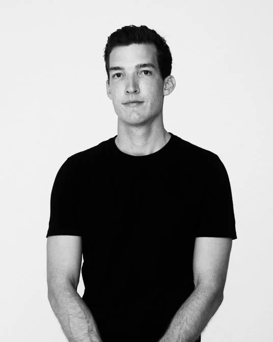 Black-and-white photo of a young man with short dark hair, wearing a black t-shirt, standing against a plain white background.