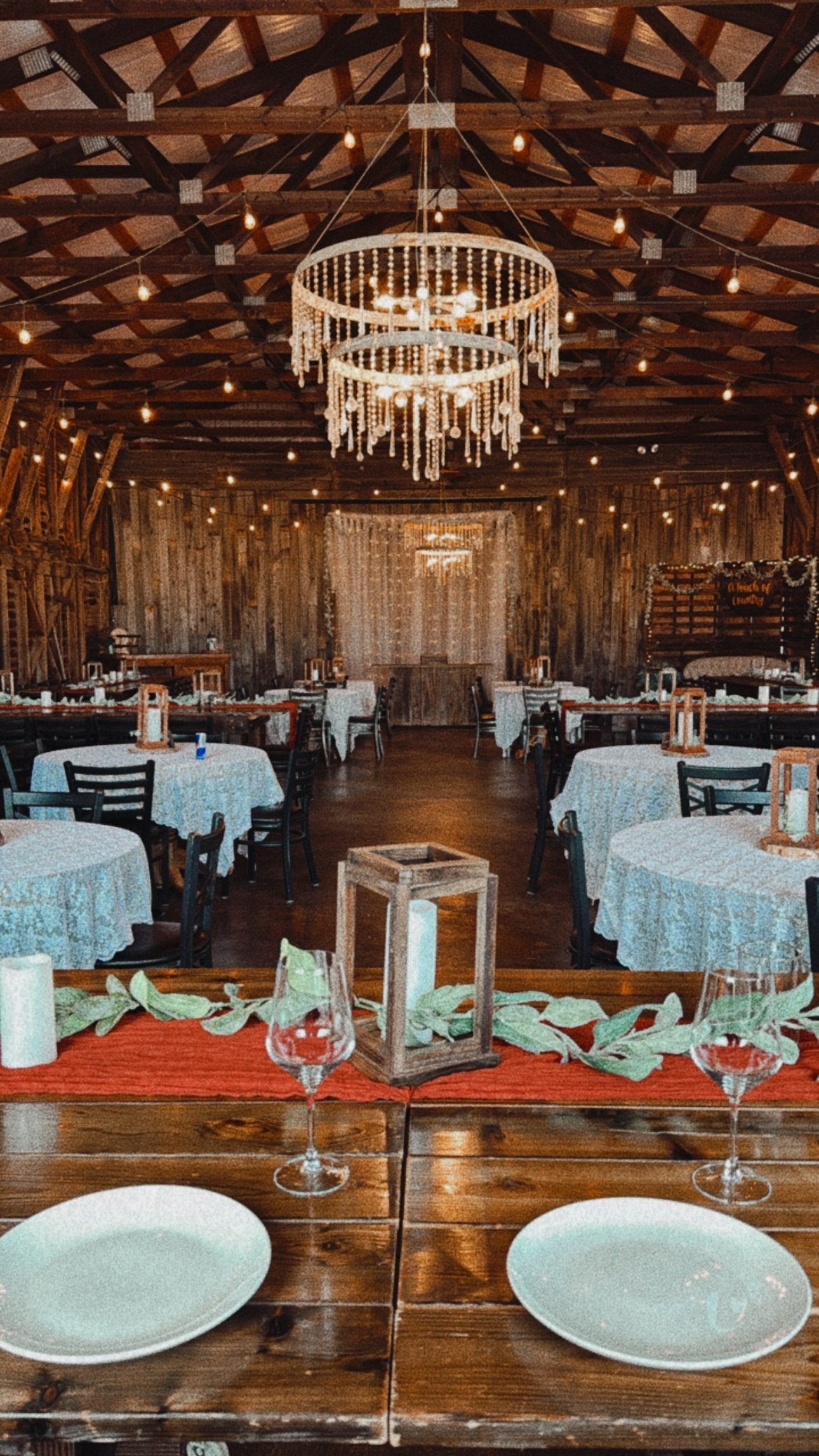 Rustic wedding reception with round tables, white tablecloths, and wooden chairs, decorated with lanterns and candles; wooden barn interior with string lights and a chandelier.