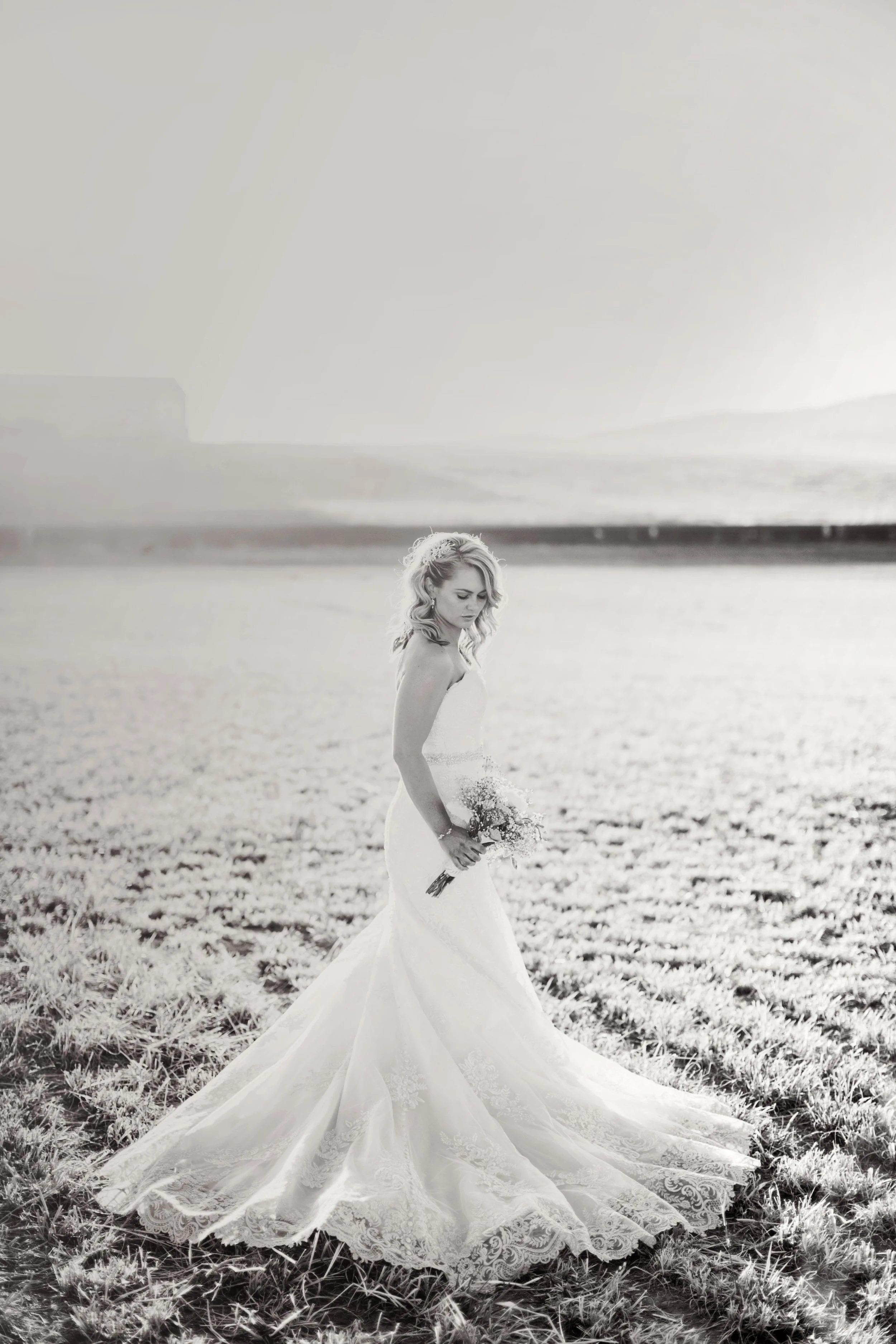 Bride in white lace dress holding bouquet in field, black and white photo.