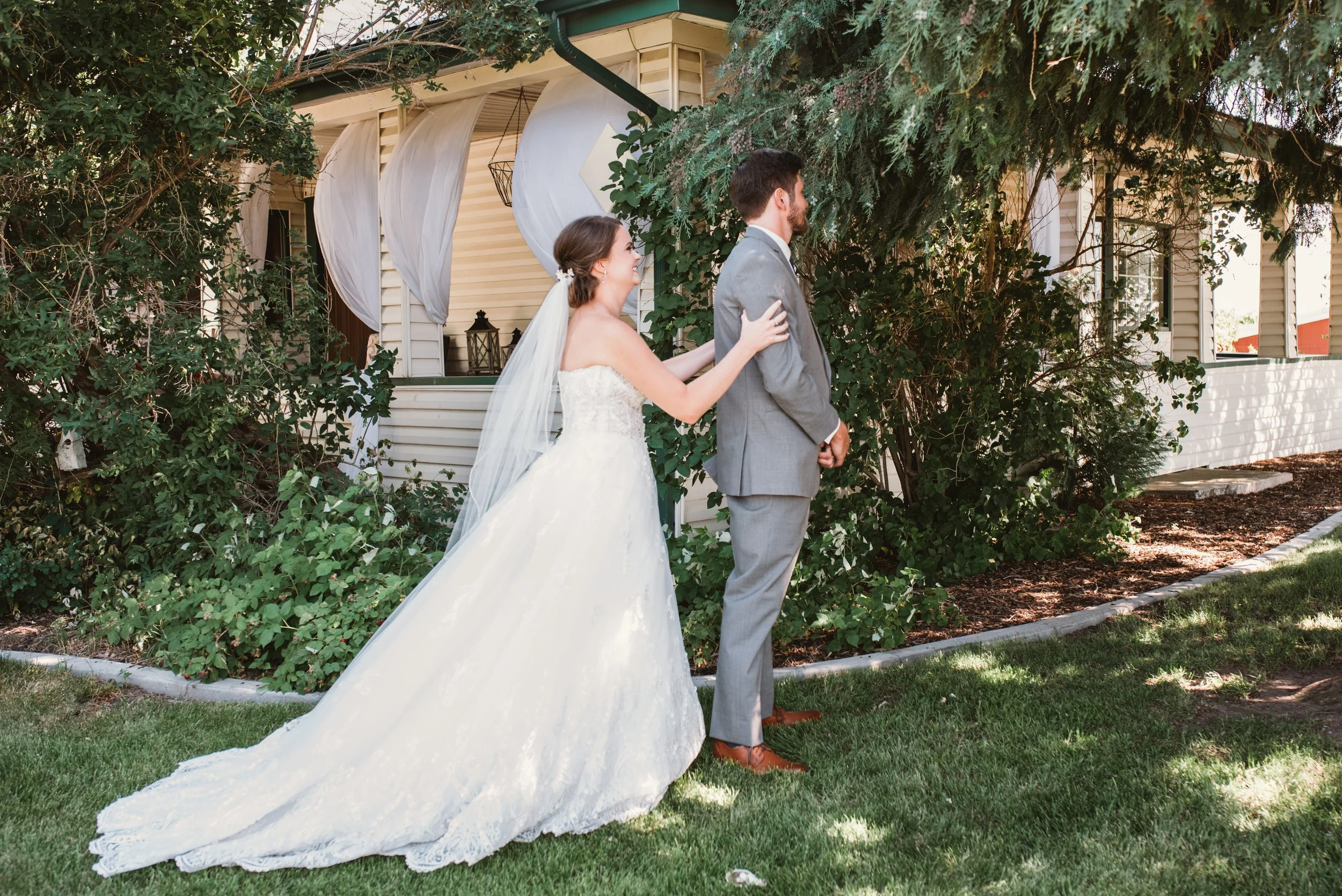 A bride in a white wedding dress approaches a groom in a gray suit from behind in a garden setting. The groom stands with his back turned, and the bride touches his shoulder. The scene is in front of a house with greenery and white drapes.