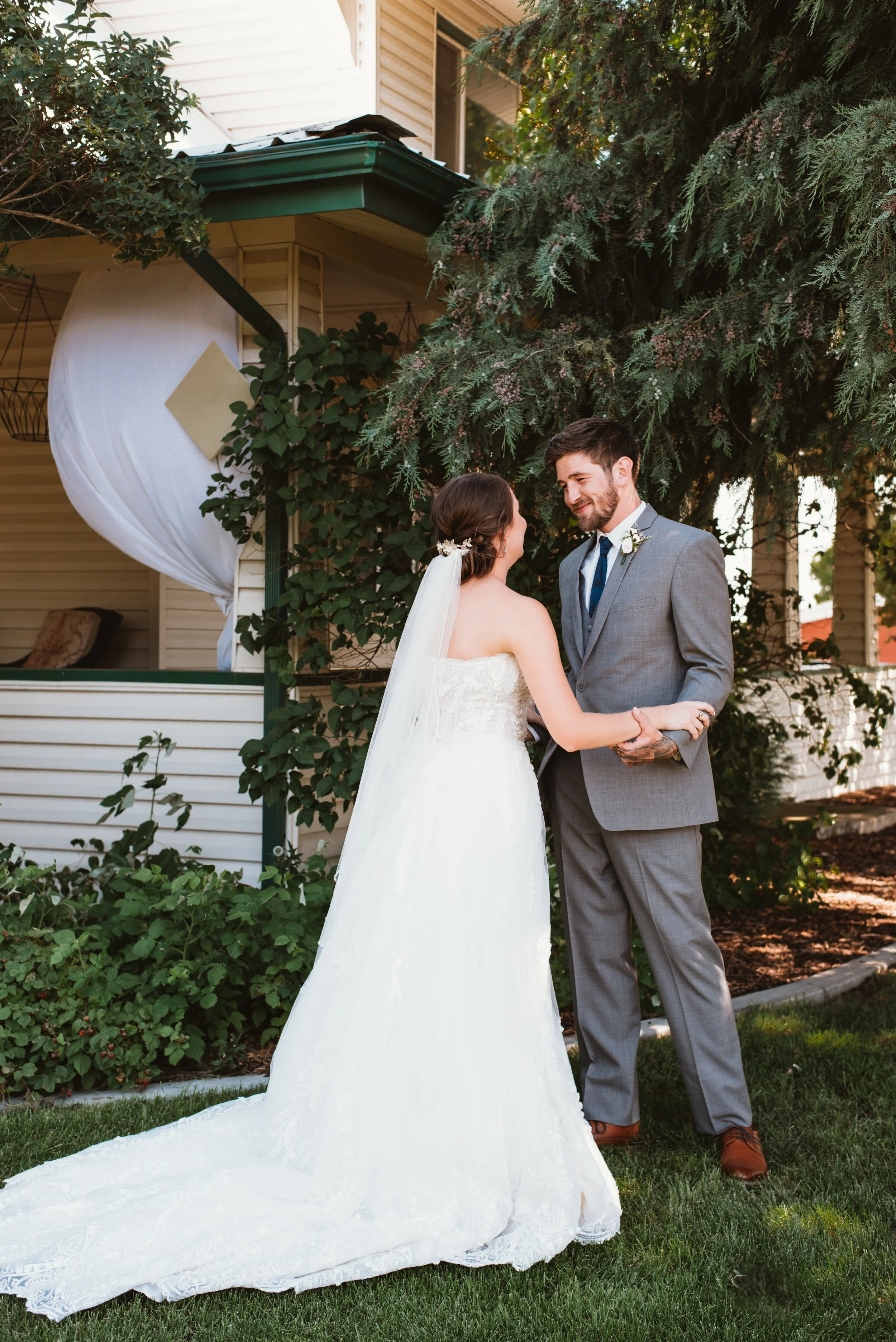 Bride in white dress and groom in gray suit holding hands outdoors near house and tree.