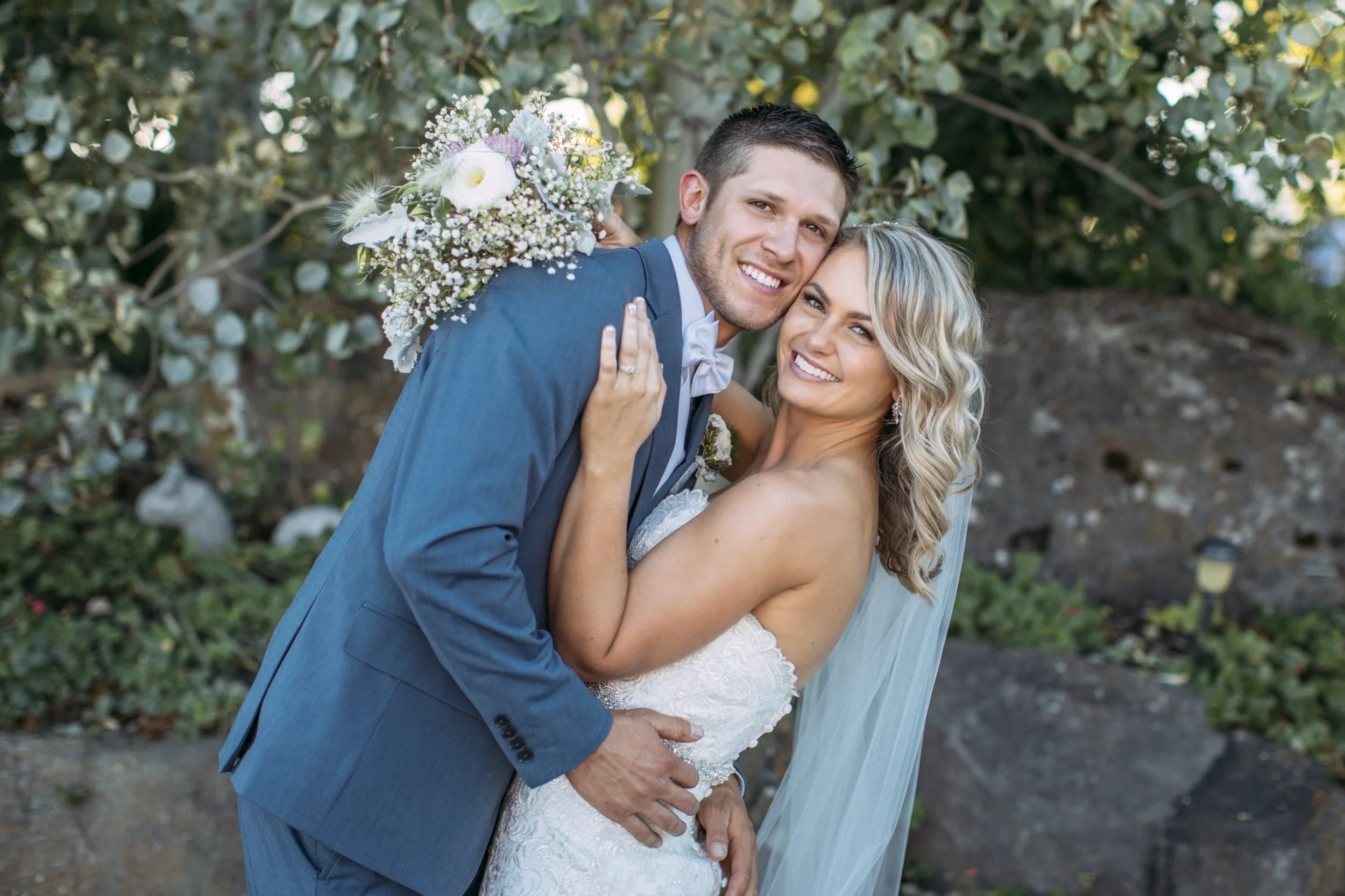 Bride and groom smiling outdoors, embracing, with greenery and rocks in background.