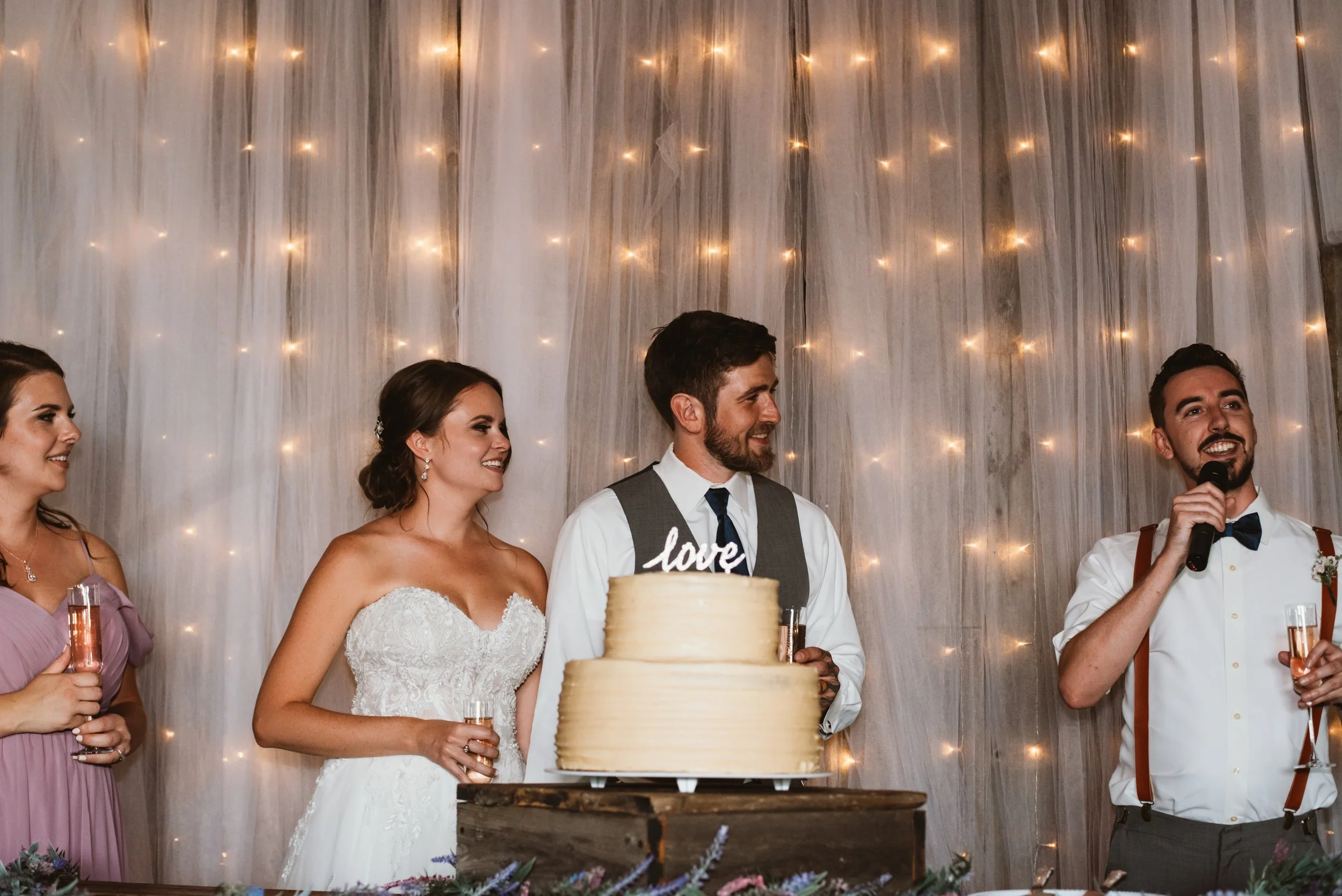 Wedding reception scene with bride and groom smiling, tiered cake with "love" topper in foreground, two guests holding champagne glasses; backdrop of twinkling lights.