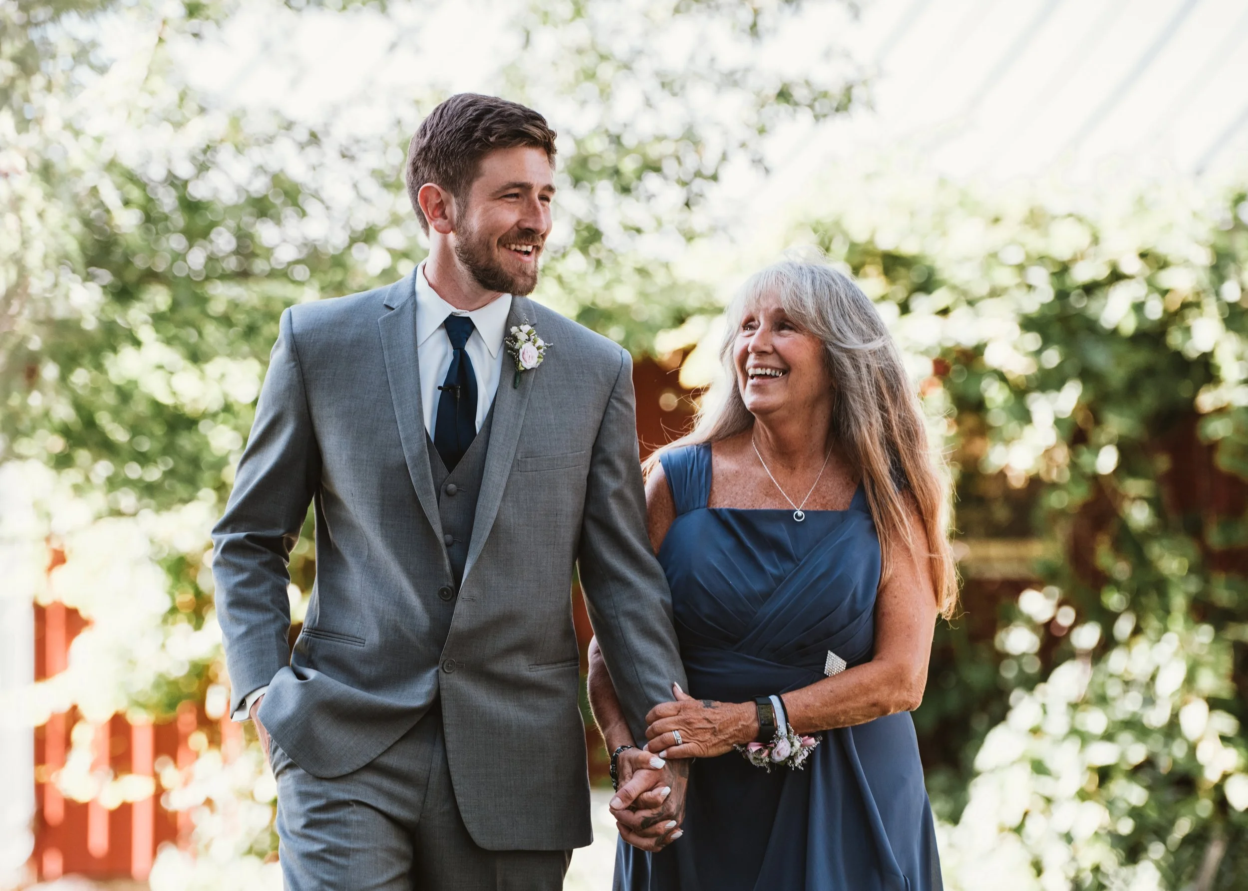 A man in a gray suit and a woman in a blue dress walking together, holding hands, and smiling outdoors with greenery in the background.