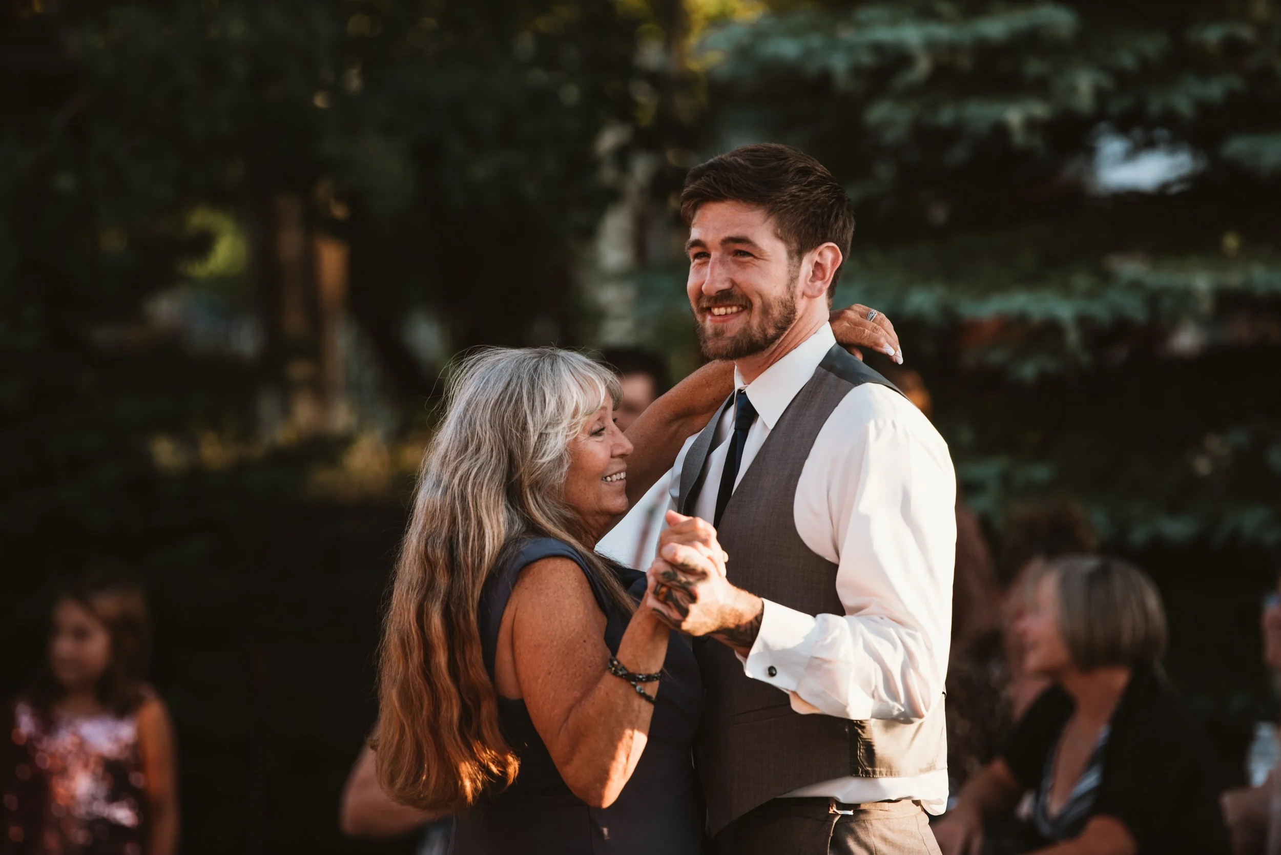 A smiling man in a suit vest dancing with a woman with long gray hair at an outdoor event, surrounded by a blurred background of trees and people.